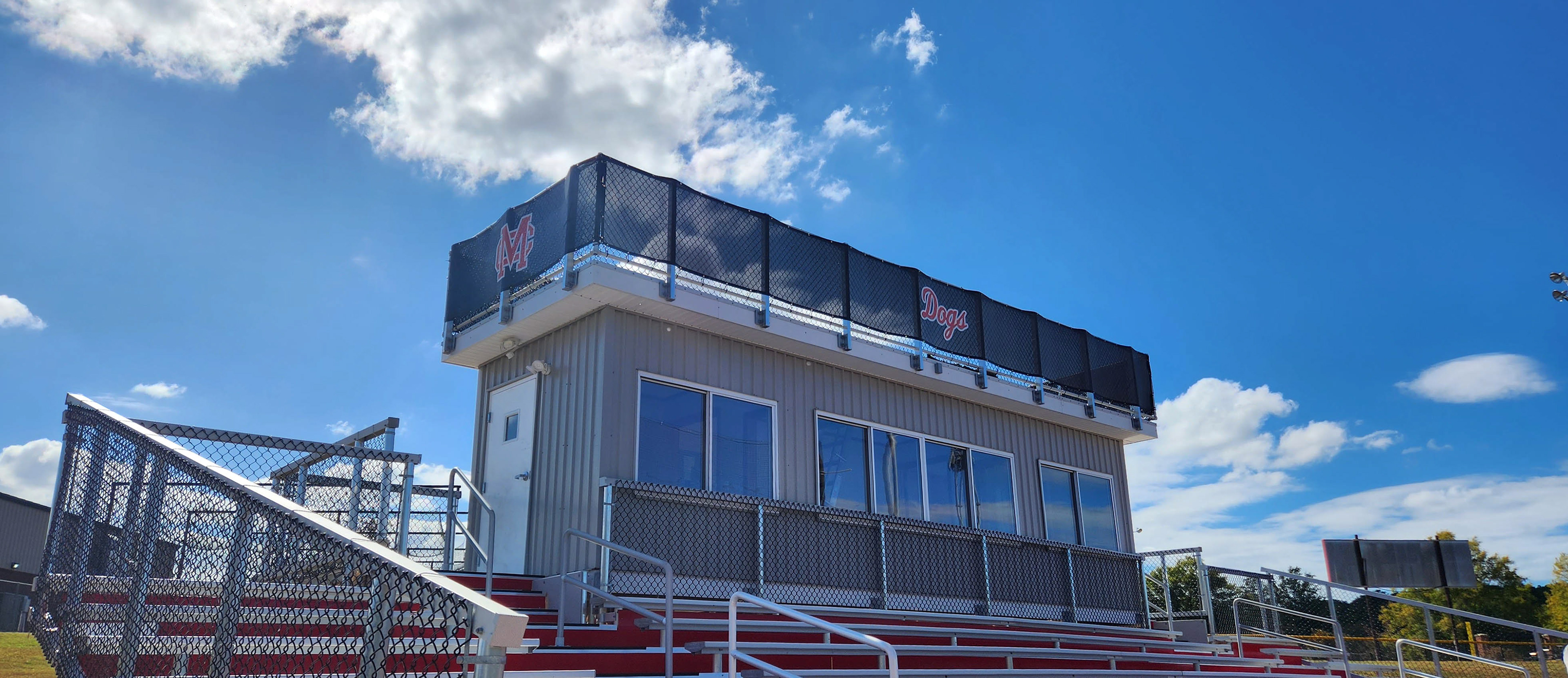 Branded black windscreen affixed to fencing, wrapping around the top of a press box and above a set of bleachers. One side is printed with the word 'Dogs' in red outlined in white, while the other side, over a door, features a red 'MC' monogram logo outlined in white.