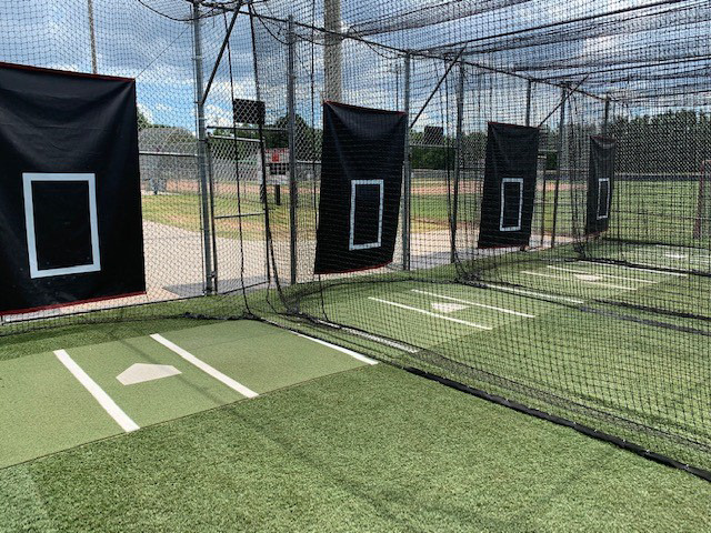 A fenced-in baseball batting cage with black sports netting separating the lanes and lining the cage. The ground is covered in green turf, and a black sports vinyl strike zone panel hangs in each lane, with a printed home plate directly in front of each panel.