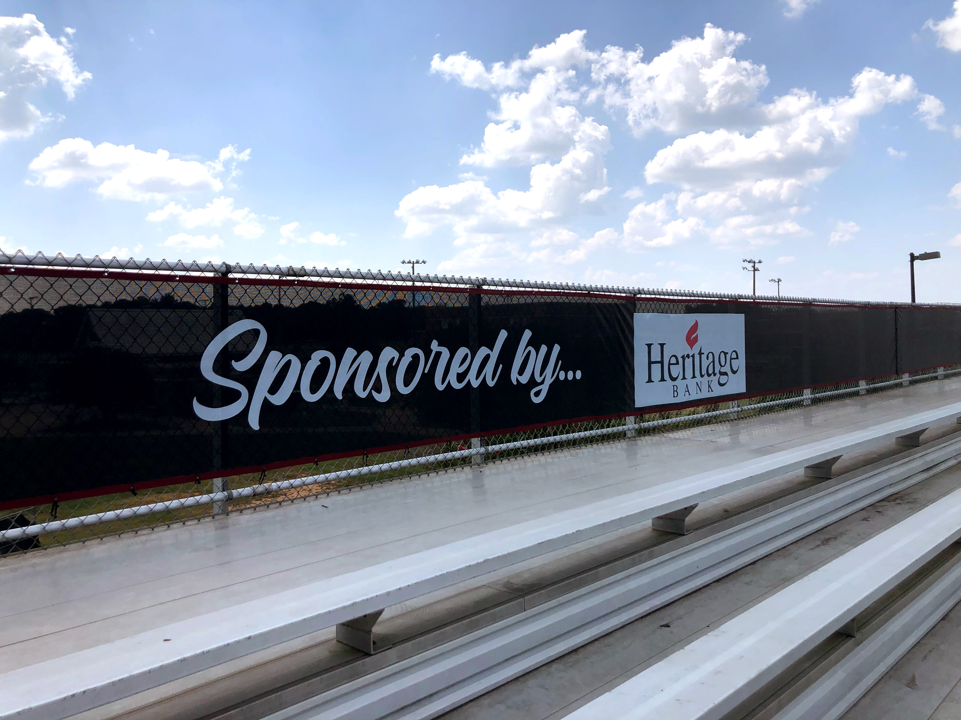 A branded windscreen panel attached to the back railing of bleachers, viewed from a slightly right angle, with clouds in the background. The windscreen features the text 'Sponsored by...' in white, 'Heritage Bank' in navy blue with a red squiggle dotting the 'i', on a white rectangular background.