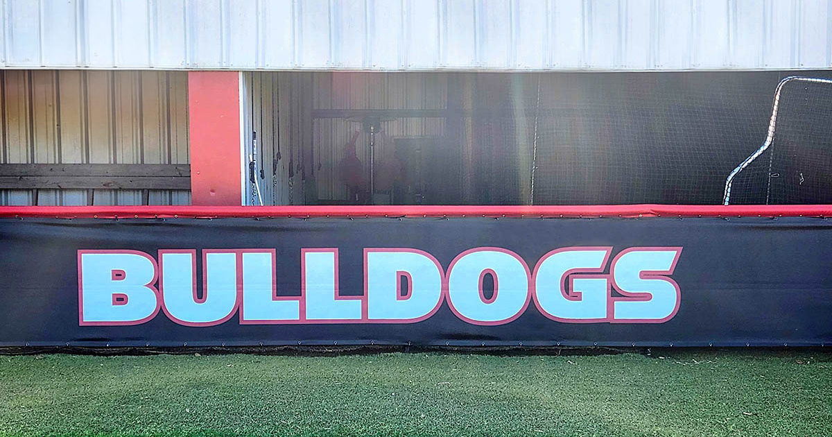 Black fence windscreen for a dugout, featuring the word 'Bulldogs' printed in white with a red outline. The windscreen is attached to a fence topped with red vinyl-covered round dugout rail padding.