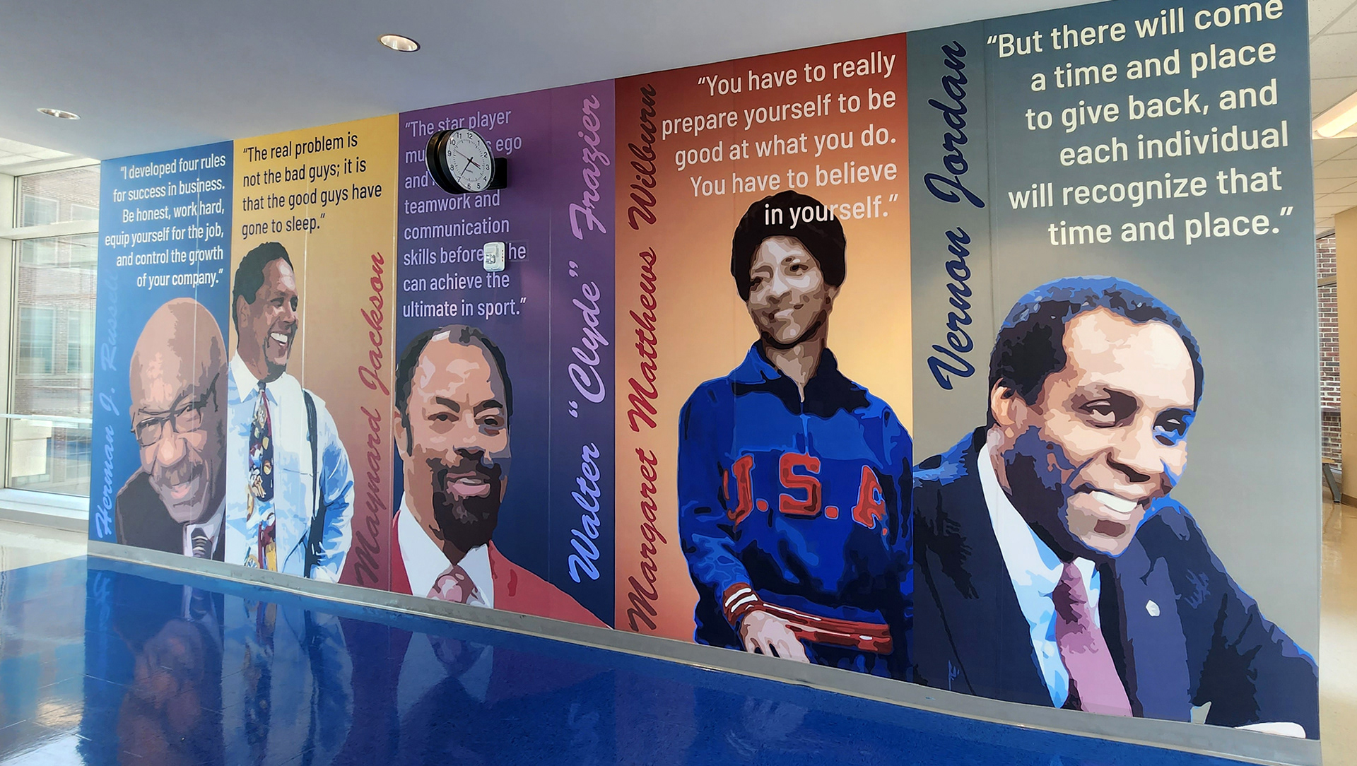 A floor-to-ceiling adhesive vinyl wall mural in a hallway. The mural features quotes and images from various individuals, each with a different color background for their rectangular space. From left to right: The first section has white text on a blue background, reading 'I developed four rules for success in business. Be honest, work hard, equip yourself for the job, and control the growth of your company' Above an image of Herman J. Russel, with his name to the side in black. The second section features black text on a gradient yellow to red background, reading 'The real problem is not the bad guys; it is that the good guys have gone to sleep.' Above an image of Maynard Jackson, with his name to the side in black. The third section has lilac text on a darker purple background, reading 'The star player must slay his ego and develop teamwork and communication skills before they can achieve the ultimate in sports' Above an image of Walter "Clyde" Frazier, with his name to the side in lilac. The fourth section has light oragne text on a gradient red and orange background, reading 'You have to really prepare yourself to be good at what you do' Above an image of Margaret Matthews Wilburn, with her name to the side in a dark red orange. The final section features light yellow text on a gray background, reading 'But there will come a time and place to give back, and each individual will recognize that time and place'  above an image of Vernon Jordan with his name to the side in dark blue.