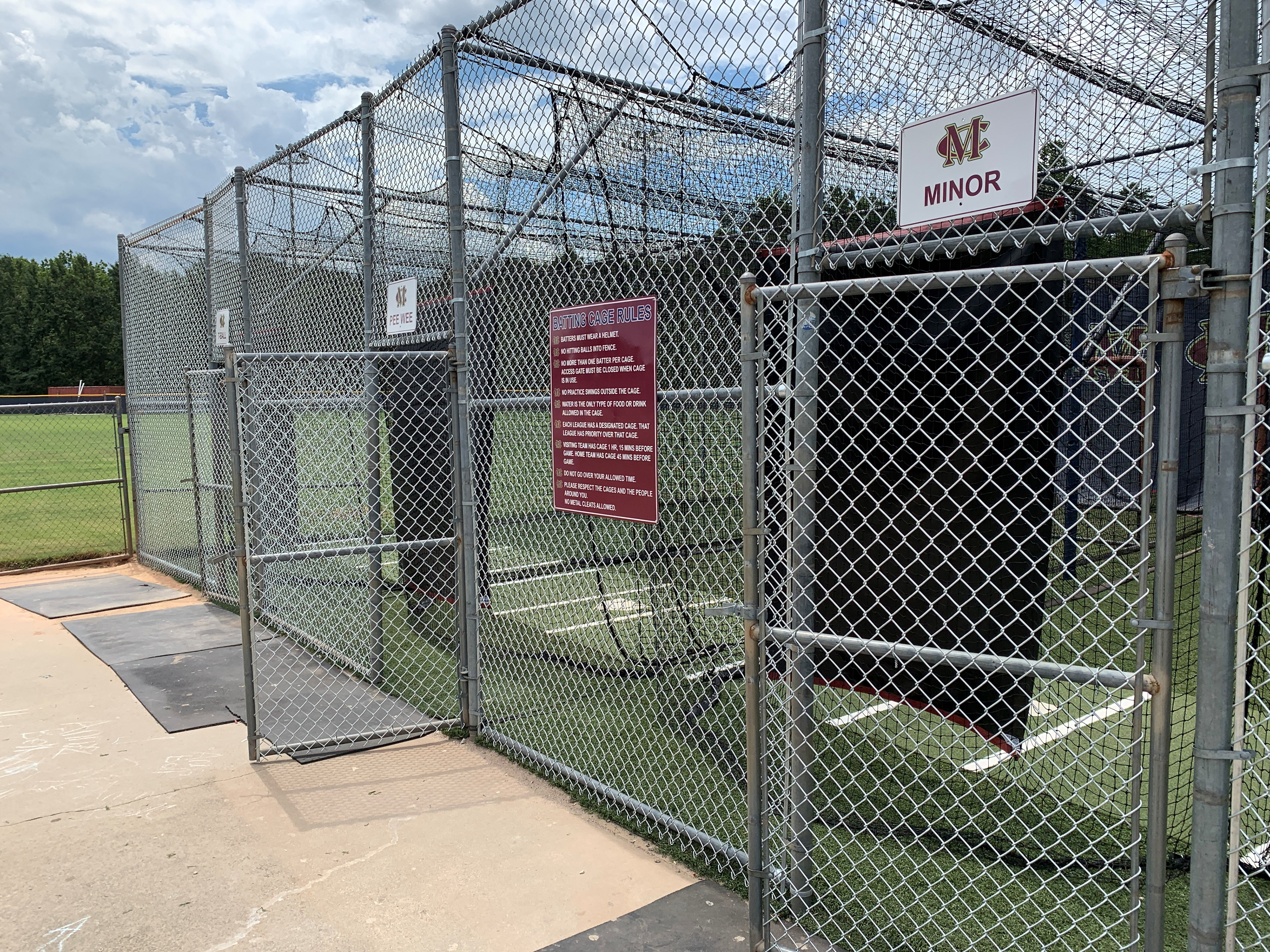 To the right a view of a silvertone metal baseball batting cage from the outside, with black sports netting separating the lanes and lining the cage, and green turf covering the ground. To the left in the background, part of a sports field is visible.