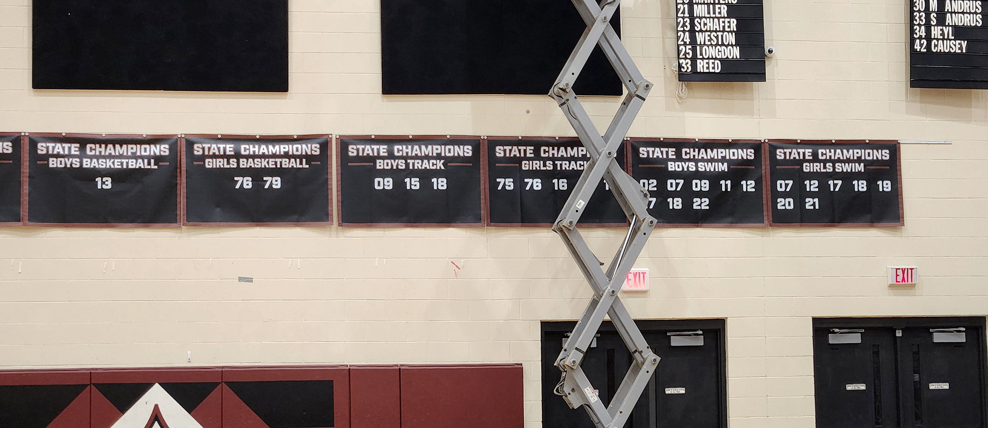 A black 10oz VCP windscren rectangular banner with a maroon border, white text and numbers promoting state champions for Wando High School. The banner is displayed on the upper part of the gym walls, just above the door