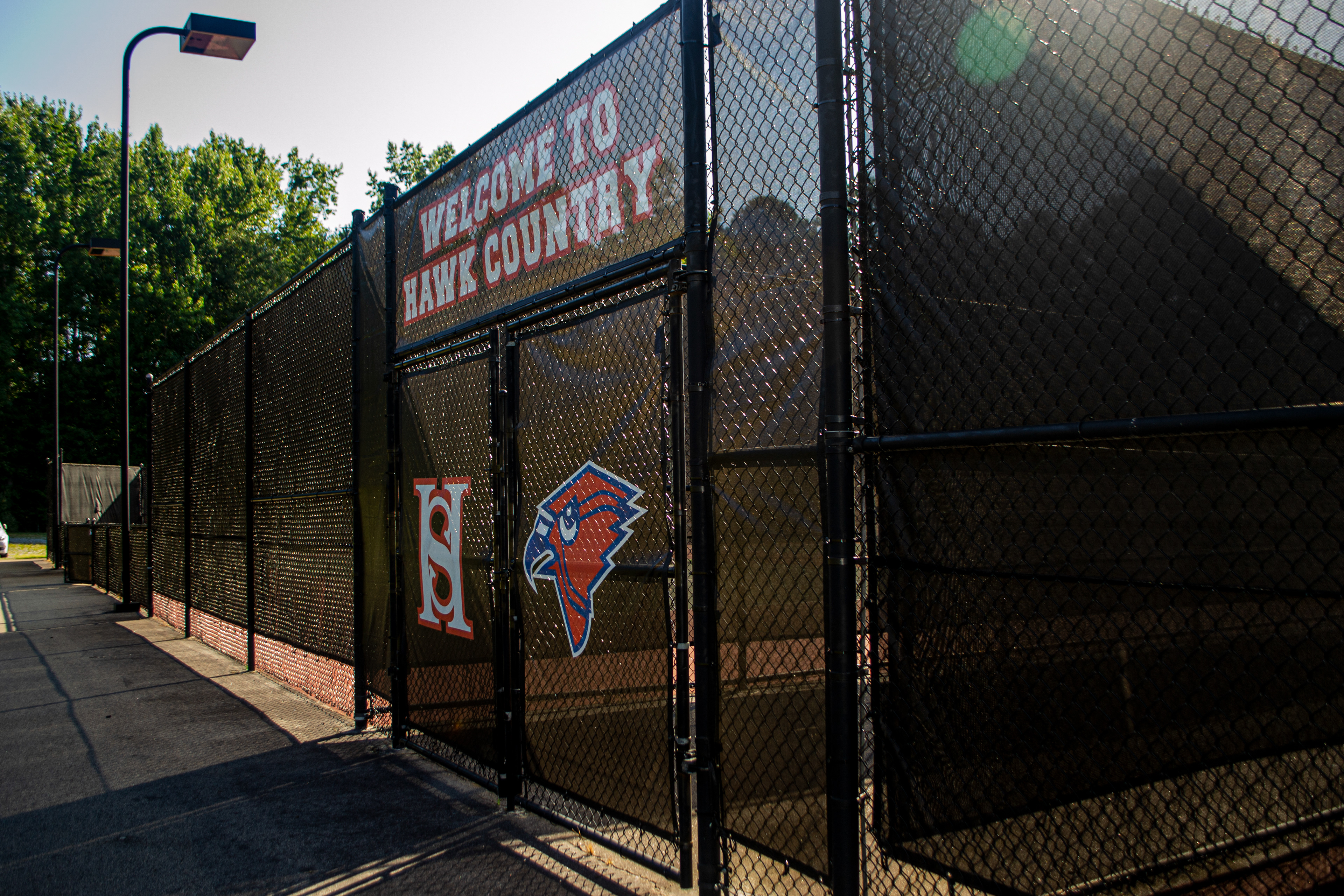A fence surrounding a tennis court viewed from the outside, with a branded black windscreen attached to the outside. Above the entrance, the windscreen displays the words 'WELCOME TO HAWK COUNTY' in white, outlined in red. Below, on the door to the left, is a white 'HS' monogram logo outlined in red. On the door to the right is a red cardinal head logo with a blue beak, outlined in blue, and then outlined again in white.