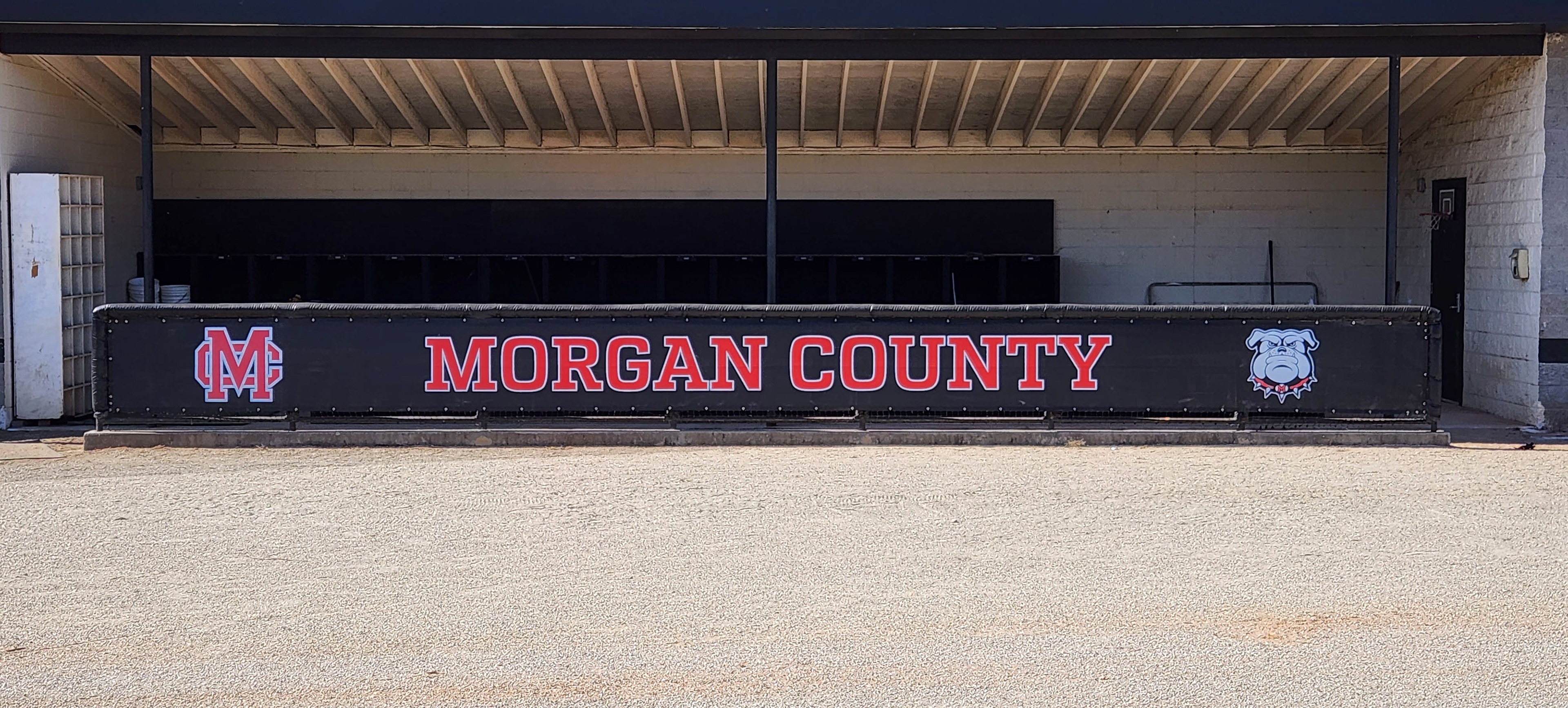 Black windscreen printed with a design from left to right: a red 'MC' logo outlined in white, the words 'MORGAN COUNTY' in red also outlined in white, and a gray bulldog logo outlined in black and white. The design is affixed to the front dugout barrier.