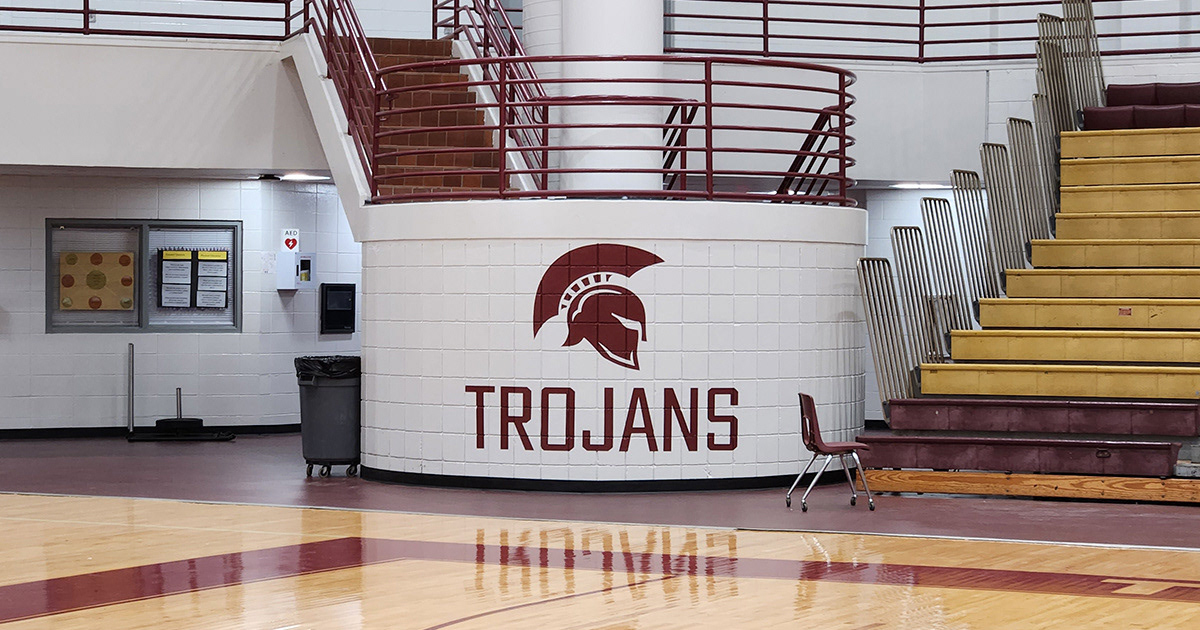 Adhesive vinyl lettering reading "TROJANS," with an image of a Trojan helmet above it, located on the outside of a brick stair landing at the far end of an indoor basketball court, between the bleachers and near the concessions.