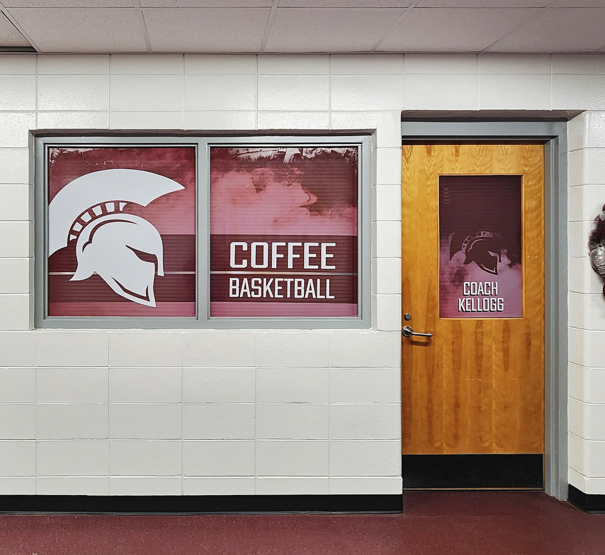 A hallway in a school, with a door on the right and a window on the left leading to the high school basketball coach's office. The left window features a full-print window perf with a burgundy background and white lettering that says 'COFFEE BASKETBALL' next to an image of a white Trojan helmet silhouette. On the top center of the door to the right is another window perf print with a burgundy smoke background, a darker burgundy Trojan helmet silhouette, and white text reading 'COACH KELLOGG".
