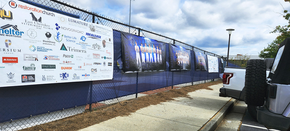A fence with a navy blue windscreen and full-print vinyl banners separates the sports area from the surrounding parking lot. The five vinyl banners include white ones on the far left and right, displaying sponsor names and logos, and three middle banners with blue and black backgrounds featuring images of players.