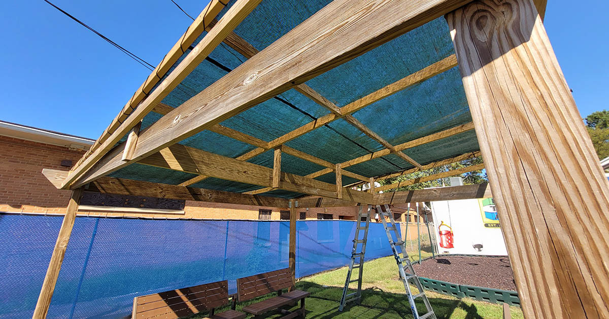 A seating area covered with a wood-framed, flat-roofed green shade structure.