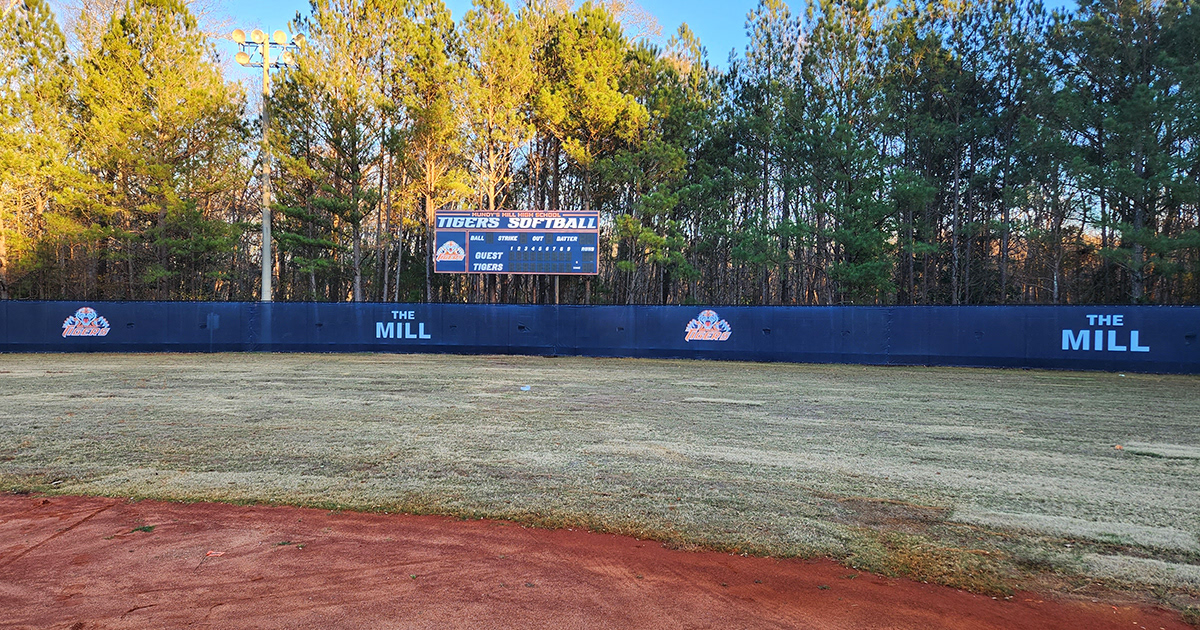 Blue fence windscreen around a baseball field with two repeating patterns: the first pattern features a tiger image above the word 'Tigers' printed in orange with a white outline, and the second pattern displays the words 'The Mill' in white. The branded windscreen matches the branded scoreboard visible just behind the fence.