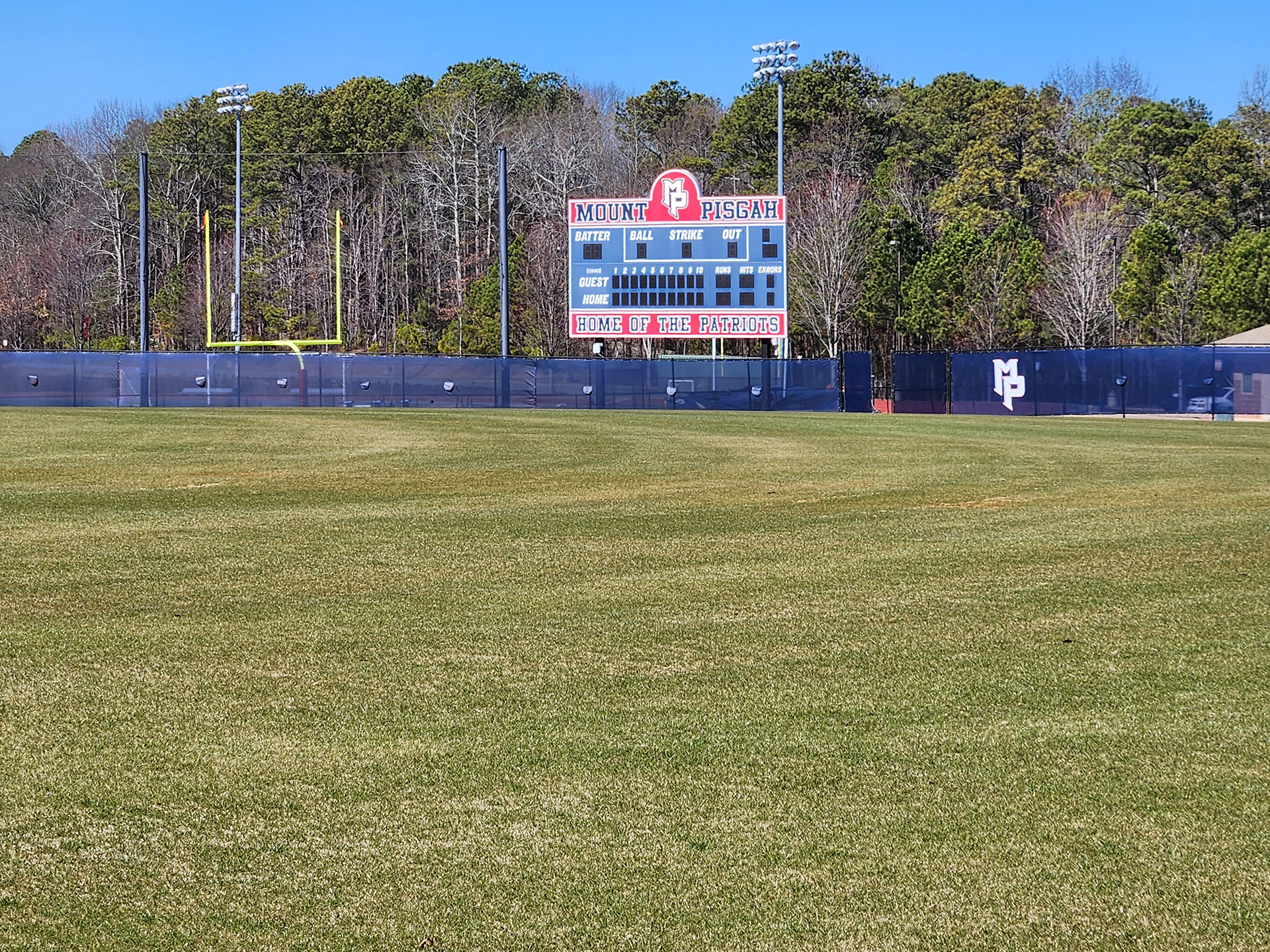 blue fence windscreen with a white logo accented in blue with red outline reading 'MP' printed on it and half moon vents around a baseball field