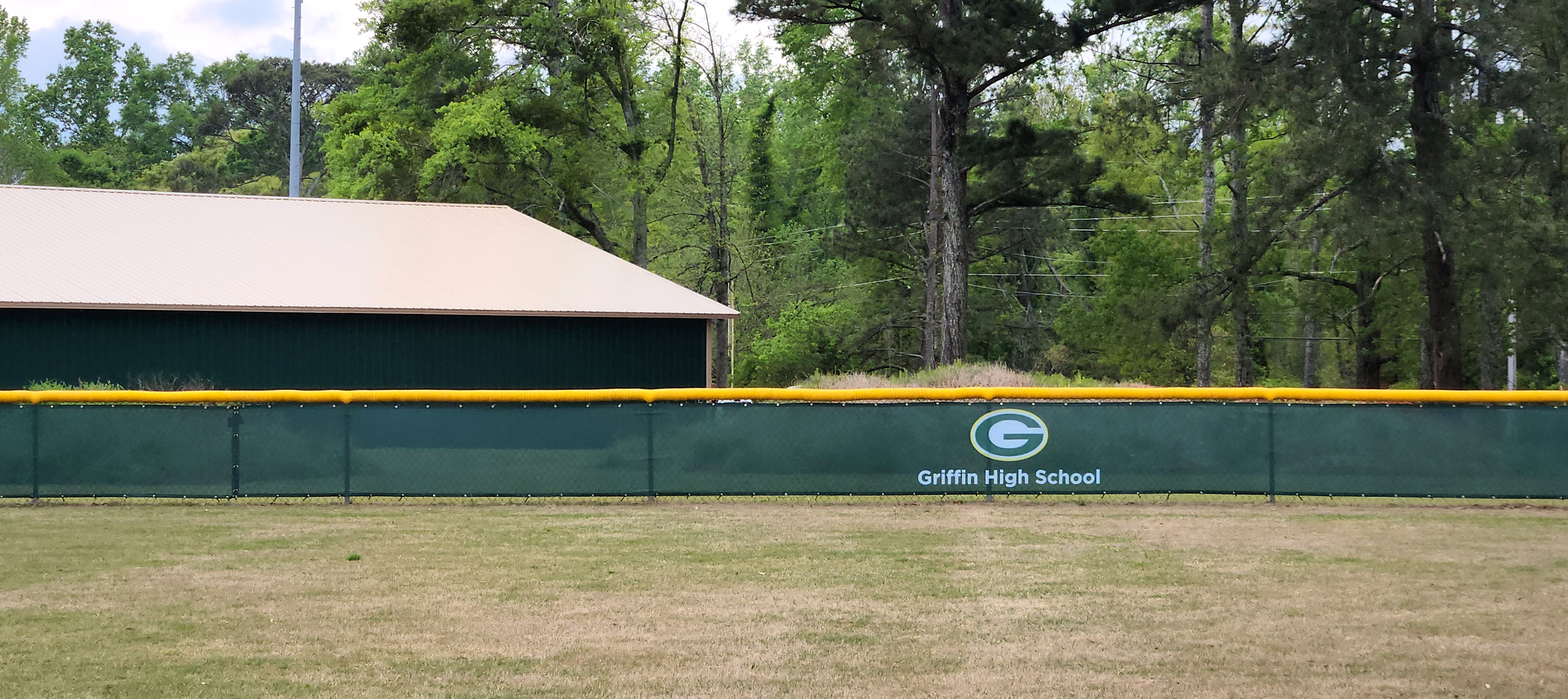 Near shot of dark forest green windscreens are branded with a white and green 'G' logo and white text reading 'Griffin High School' attached to a fence with a yellow corrugated poly cap rail top fence cap.