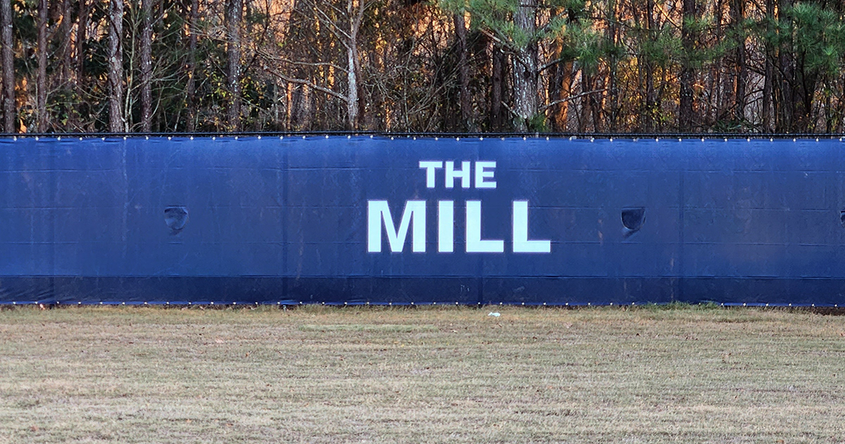 A closeup of blue fence windscreen around a baseball field with the words 'The Mill' in white.