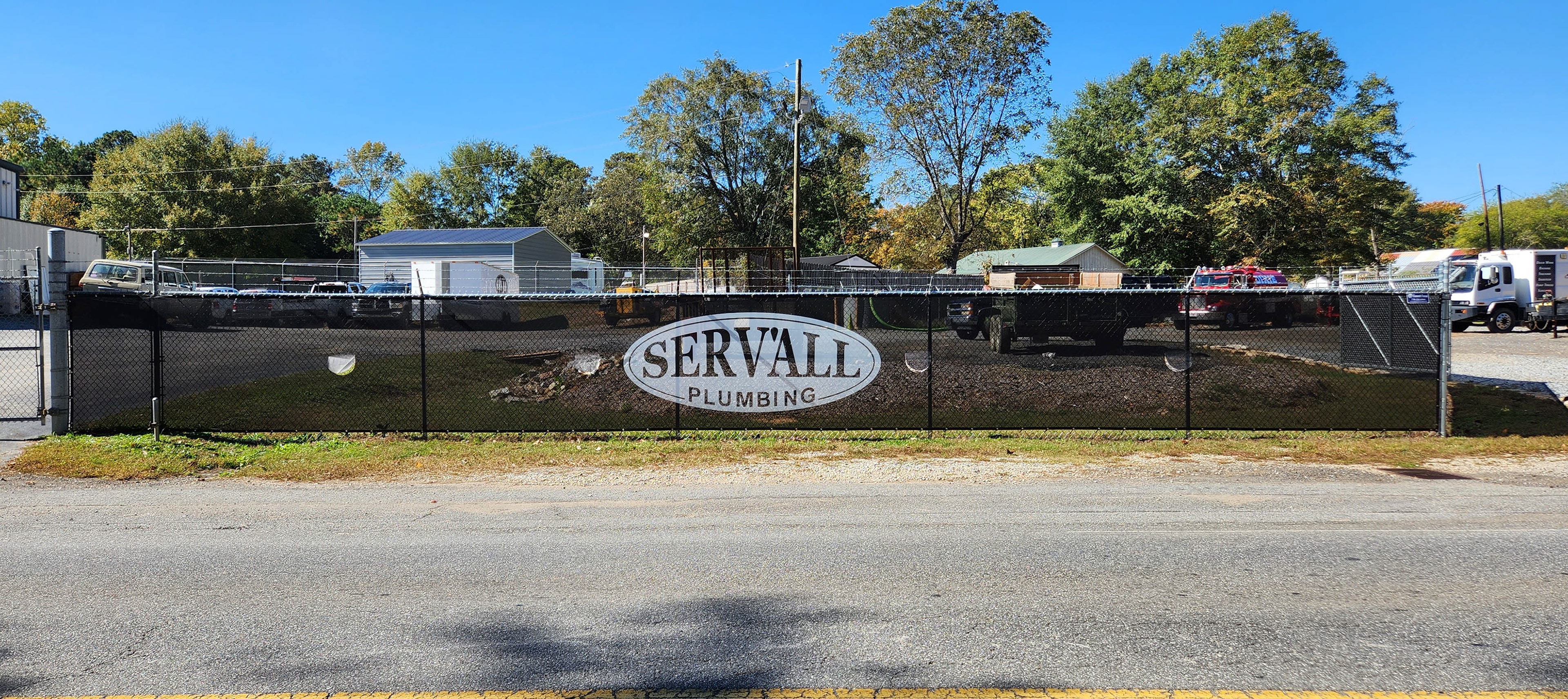 Black windscreen printed with the 'SERV'ALL PLUMBING' logo in white, affixed to a fence around a business lot.
