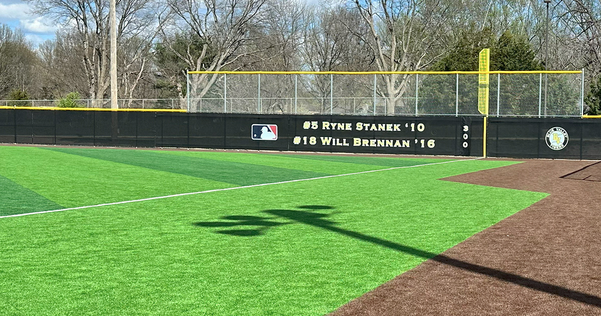 Black windscreen attached to a fence branded with an blue, red and white MLB logo next to text printed in yellowand white reading "#5 Ryne Stanek '10" and "18 Will Brennan '16". A black windscreen field marker with "300" printed in yellow and white and a black, white and yellow round logo reading "Blue Valley Tigers". Fence is topped with yellow corrugated poly cap rail top fence cap and surrounds a baseball field.
