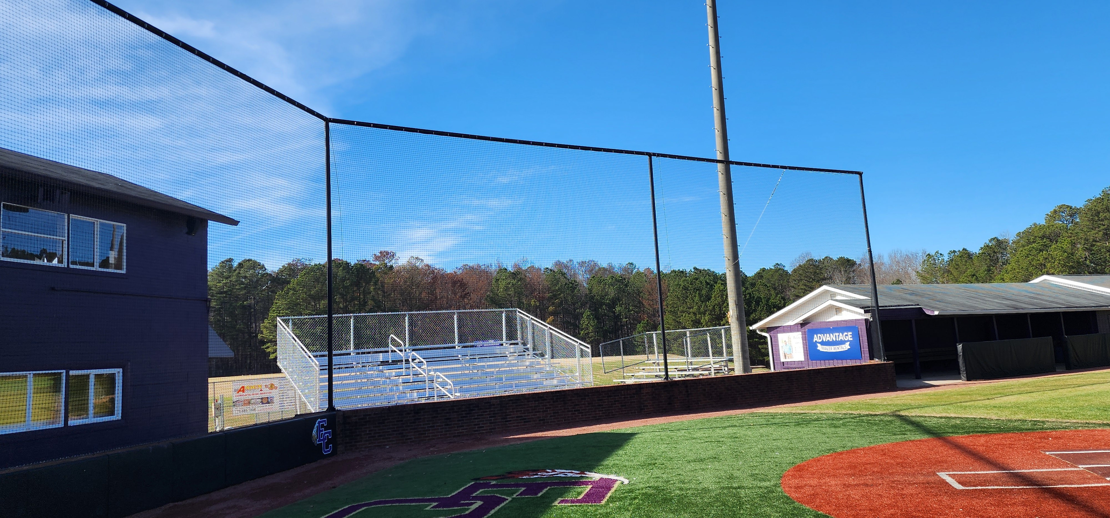backstop black sports netting surrounding the catcher's box above black sports vinyl covered backstop padding printed with blue 'EC' logo with white outline and white and yellow feather accents, attached to a brick wall