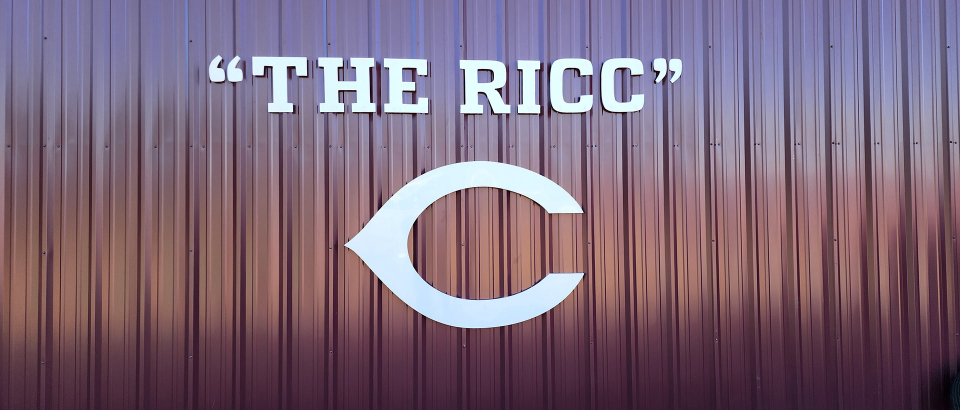 Routed plastic signage displaying the words "The Ricc" with a large letter "C" positioned underneath, mounted on the side of the Coffee High School's Riccardo Ingram hitting facility.