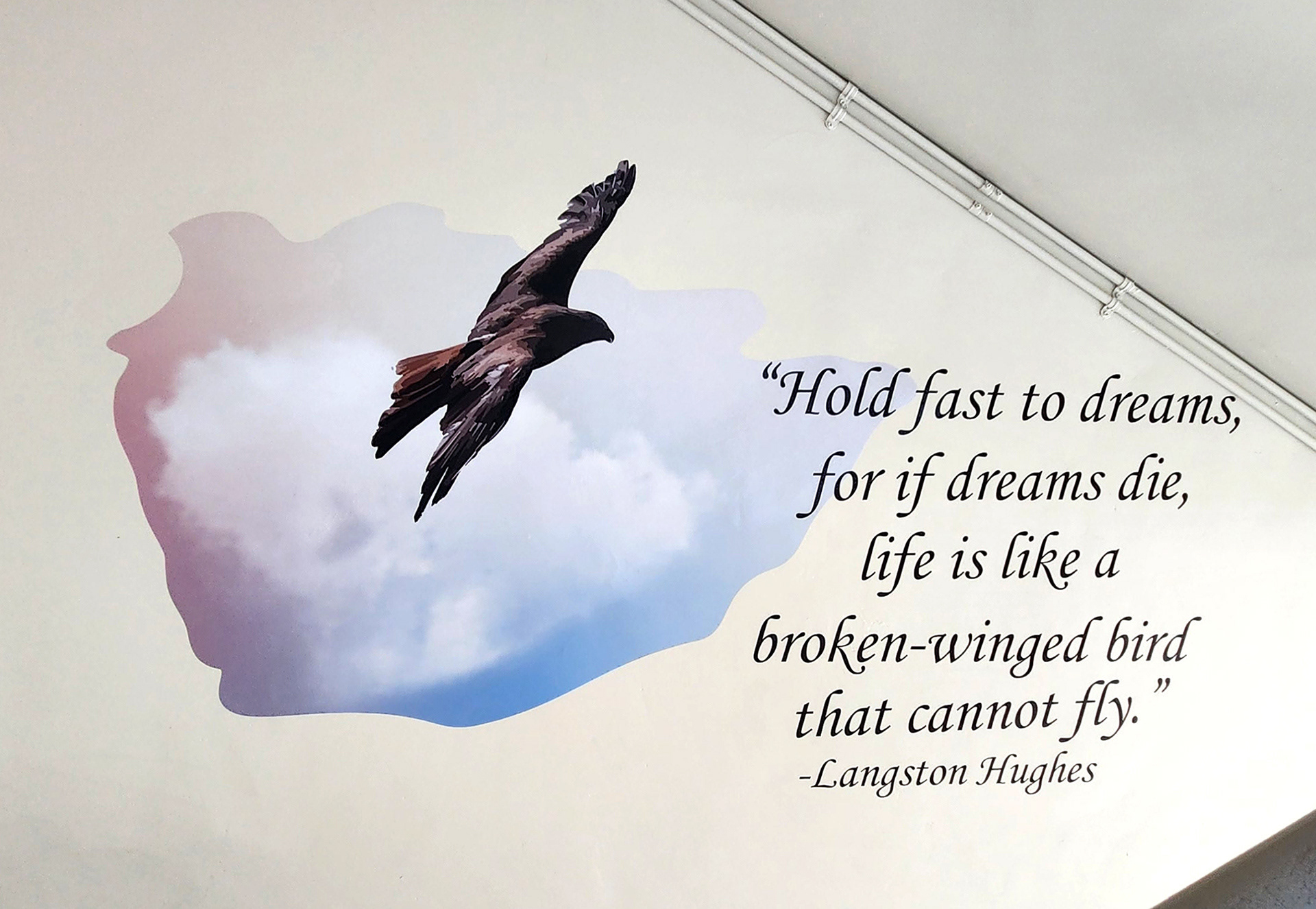On a wall near a stairwell landing or platform, an adhesive vinyl image of a black bird flying over a clouded sky, next to the quote 'Hold fast to dreams, for if dreams die, life is like a broken-winged bird that cannot fly' – Langston Hughes. 