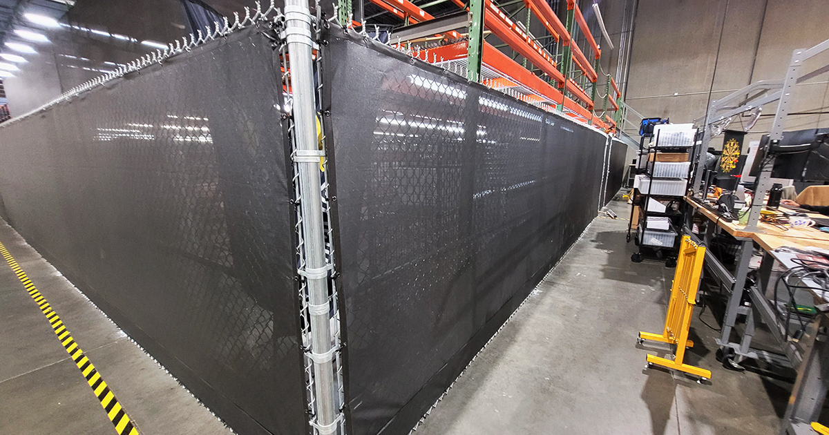 Black fence windscreen inside a warehouse, used to separate the shelving area from a bench workshop area.