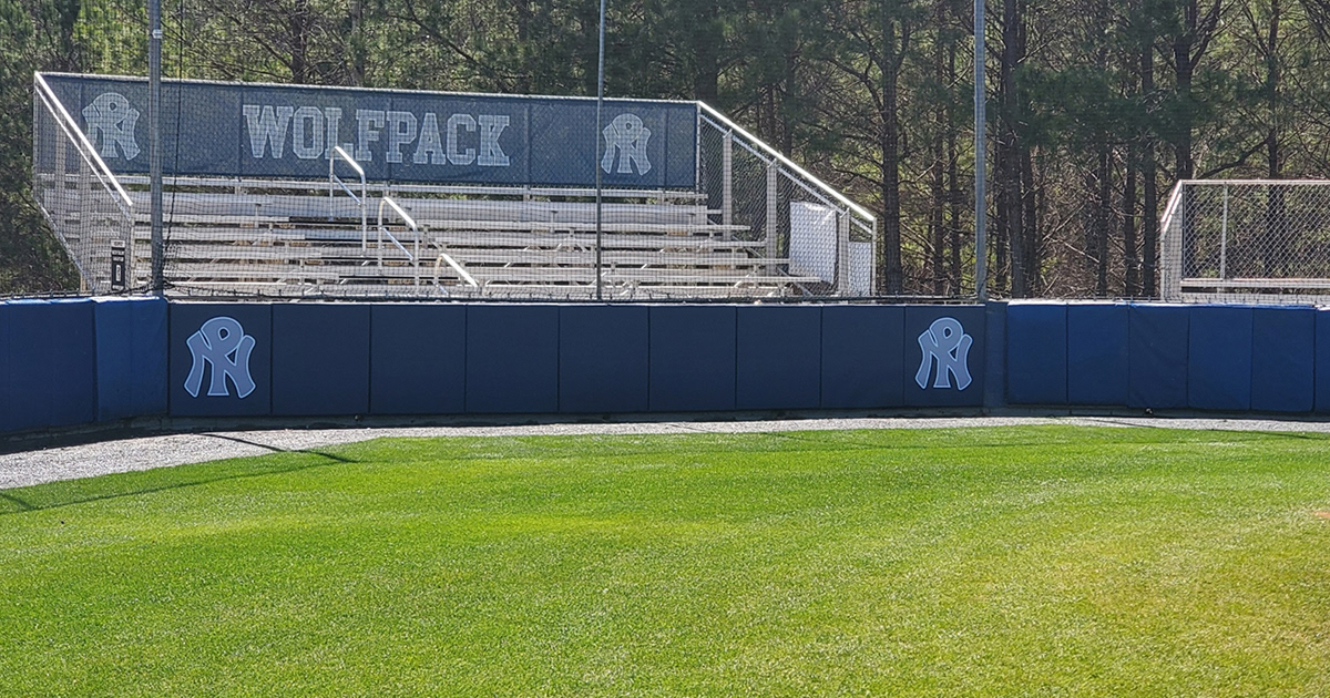 wall padding covered in navy blue sports vinyl branded with NP logo. Navy blue windscreen branded with grey lettering and logos outlined in white on the back of a bleacher stand.