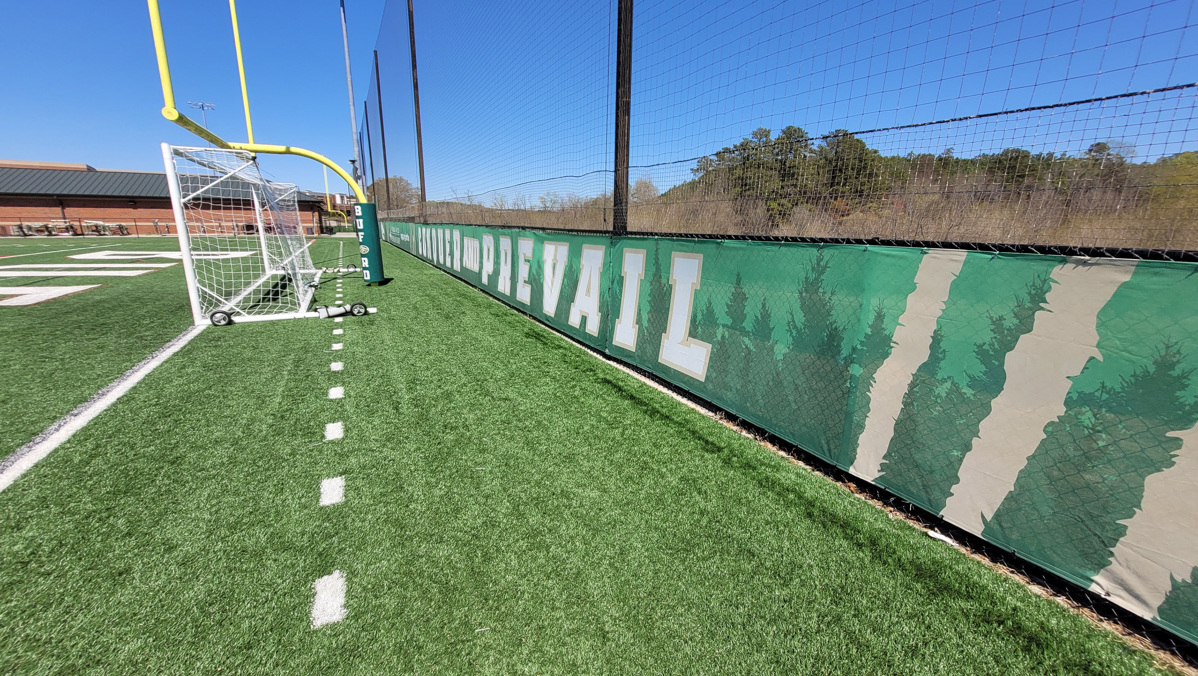 Dark evergreen tree silhouette and white lettering with a beige outline next to sponsor messages printed on a green fence windscreen at the end of a football field, near a football goal post with protective foam padding covered in branded green sports vinyl at the base, viewed from near the fence on the far right side.