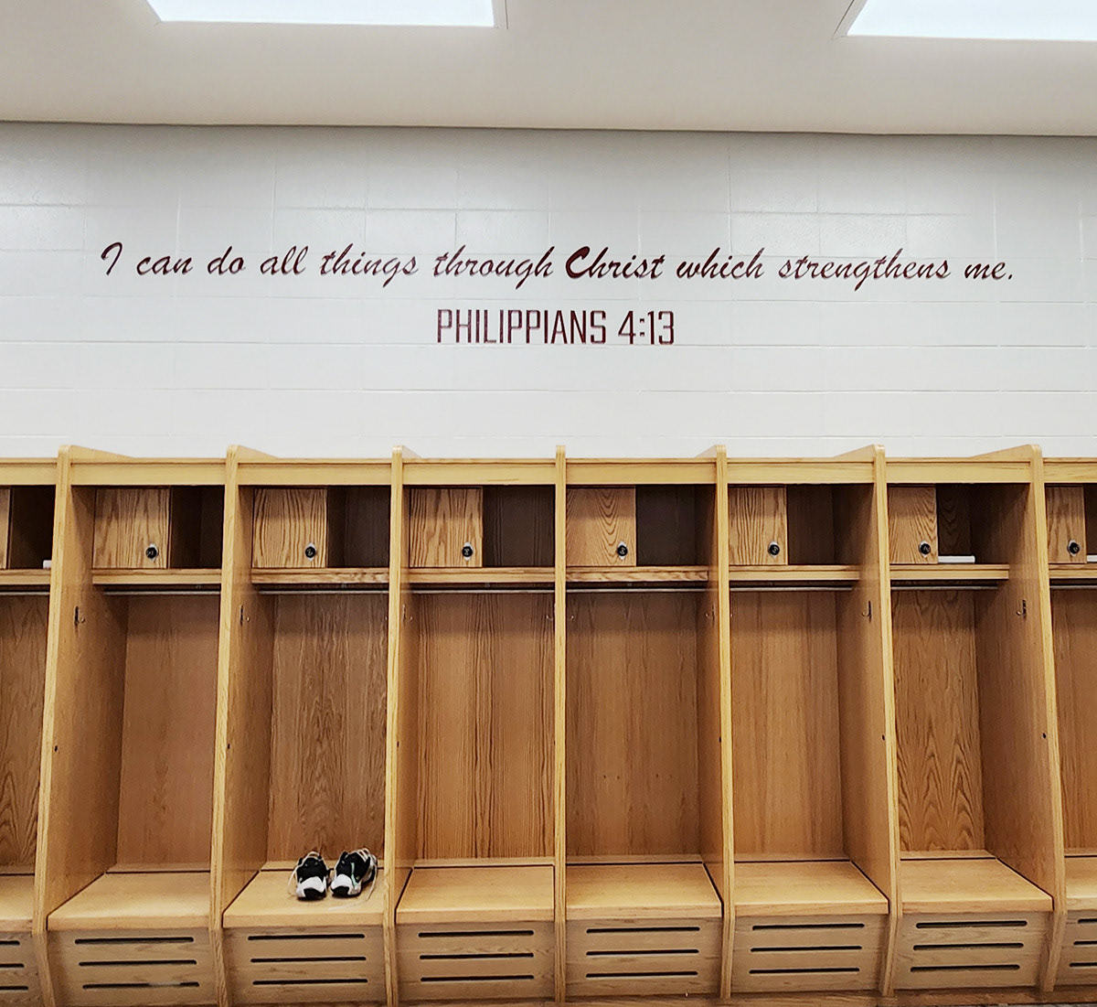 Precision-cut adhesive vinyl lettering in burgundy, displaying the Bible verse "I can do all things through Christ which strengthens me. Philippians 4:13," affixed to a painted white brick wall above wooden lockers in a high school locker room.