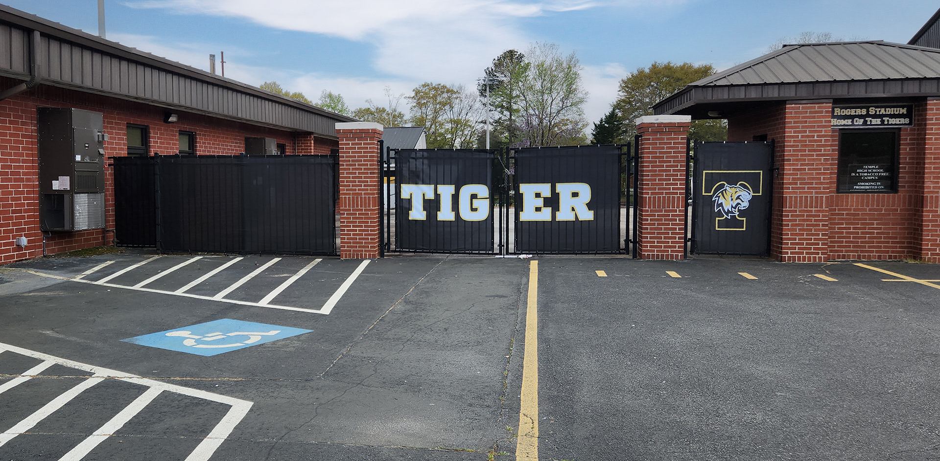 Black windscreens branded with the 'tiger nation' logo and the words "TIGER" printed in white with a yellow outline attched to fencing near two brick structures and a parking lot.