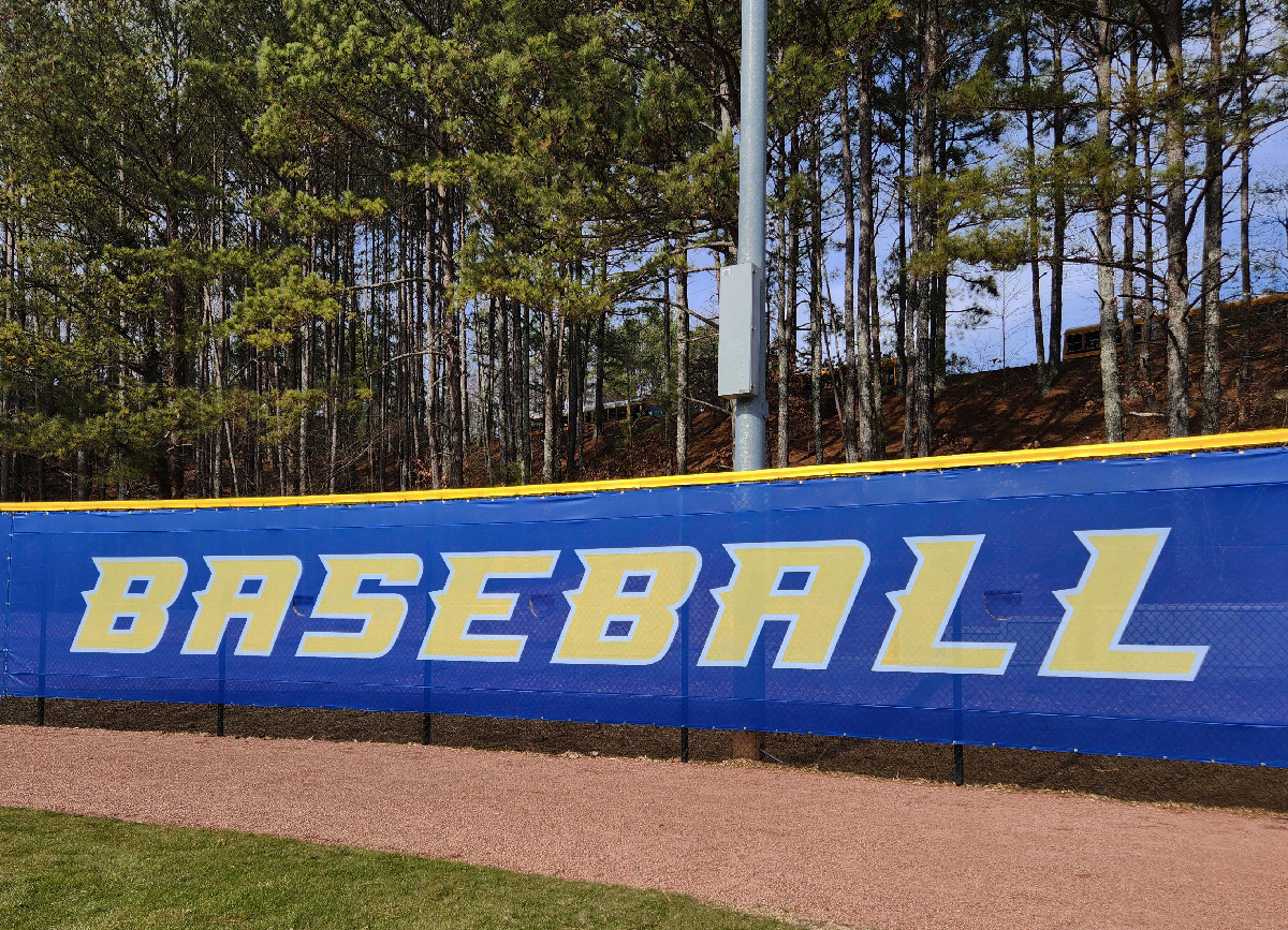 blue windscreen attached to a fence barrier topped with yellow fence guard rail top tear drop fence cap, printed with yellow text oulined in white reading 'baseball' on the outer edge of a baseball field