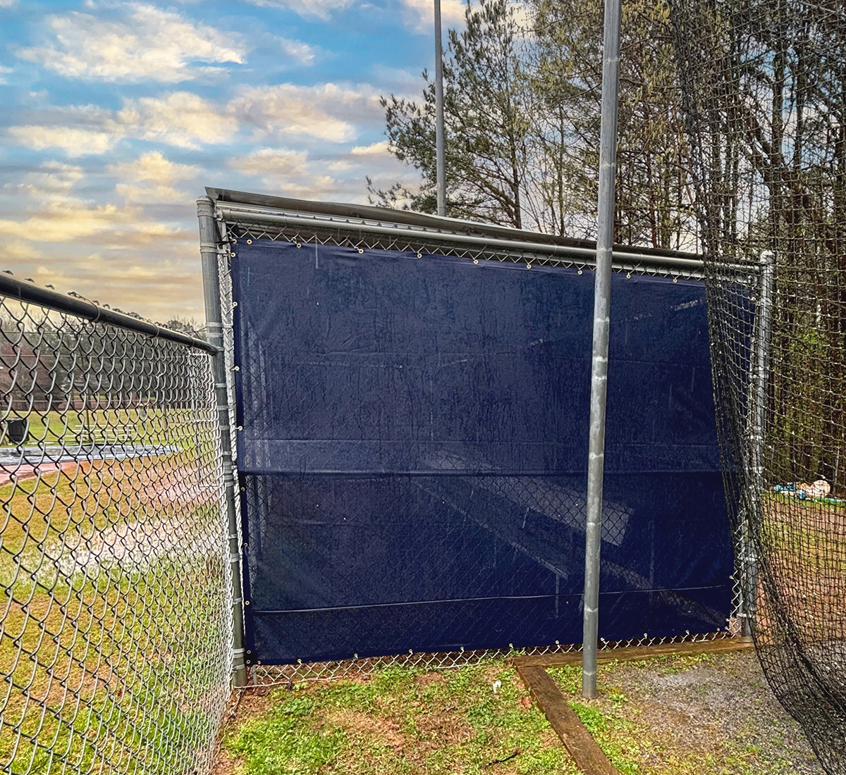 Navy blue fence windscreen branded with "MB" in white, wrapped around a baseball dugout, viewed from the left side. 