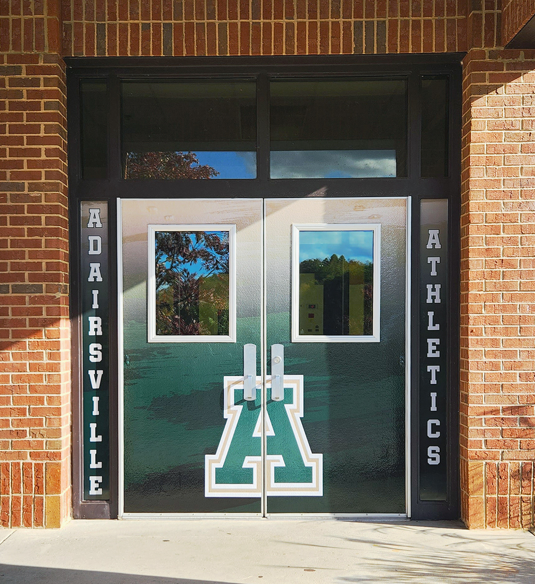 Double door entrance to a school with full print dual door wraps, creating a singular image with a gradient background and an outlined 'A' monogram in front. On either side of the doors are vertical windows with full print window perf; the left window displays the text 'ADAIRSVILLE' and the right window shows the text 'ATHLETICS'