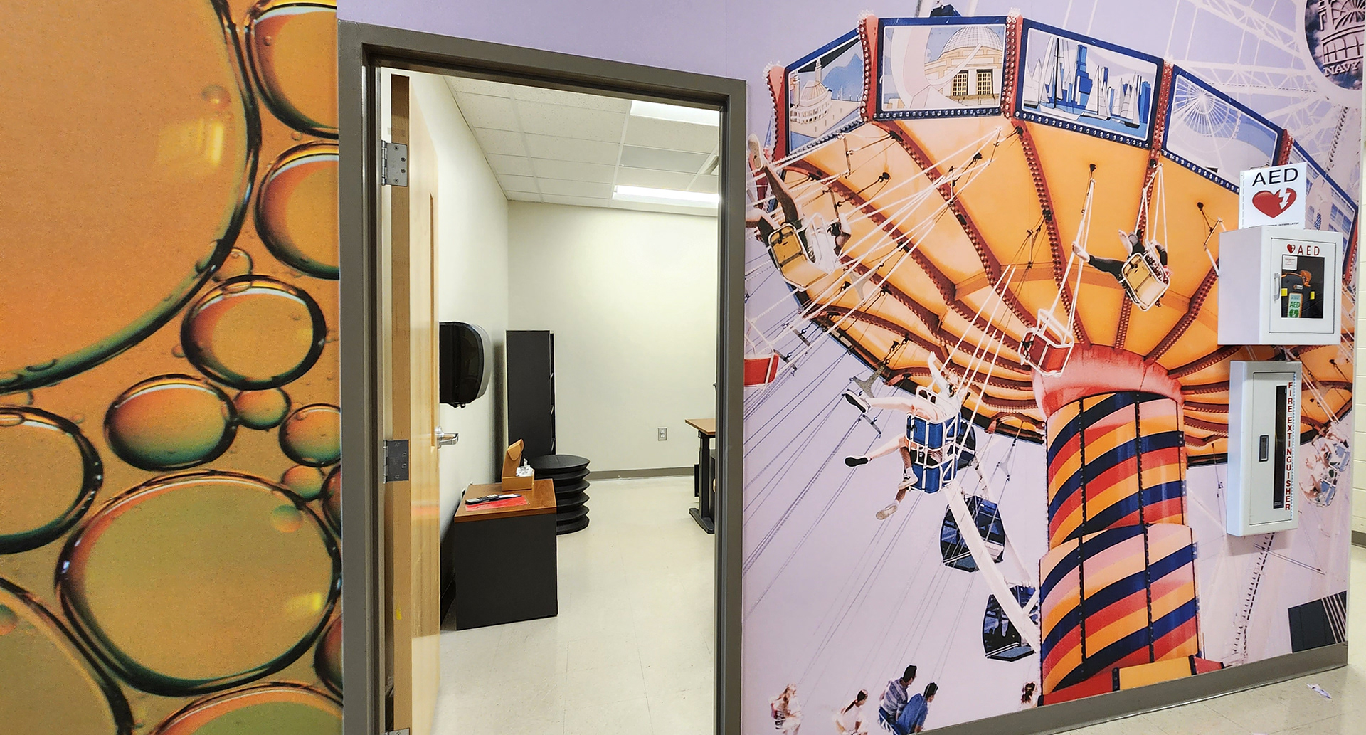 A floor-to-ceiling adhesive vinyl wall mural in a hallway, placed to the left and right of an open doorway. The mural on the left features bubbles against an orange background with green shading. The mural on the right depicts a colorful large swing ride or flying chair ride. 