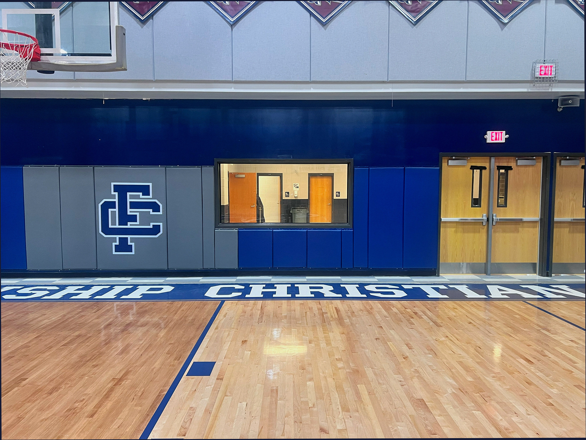 Fourteen vinyl-covered backstop pads in a gym, attached to a wall below and beside a large window. The left pads are gray, with one pad featuring a navy blue 'FC' monogram logo outlined in white. The right pads are blue and extend to the edge of the wall, ending by a set of double doors.