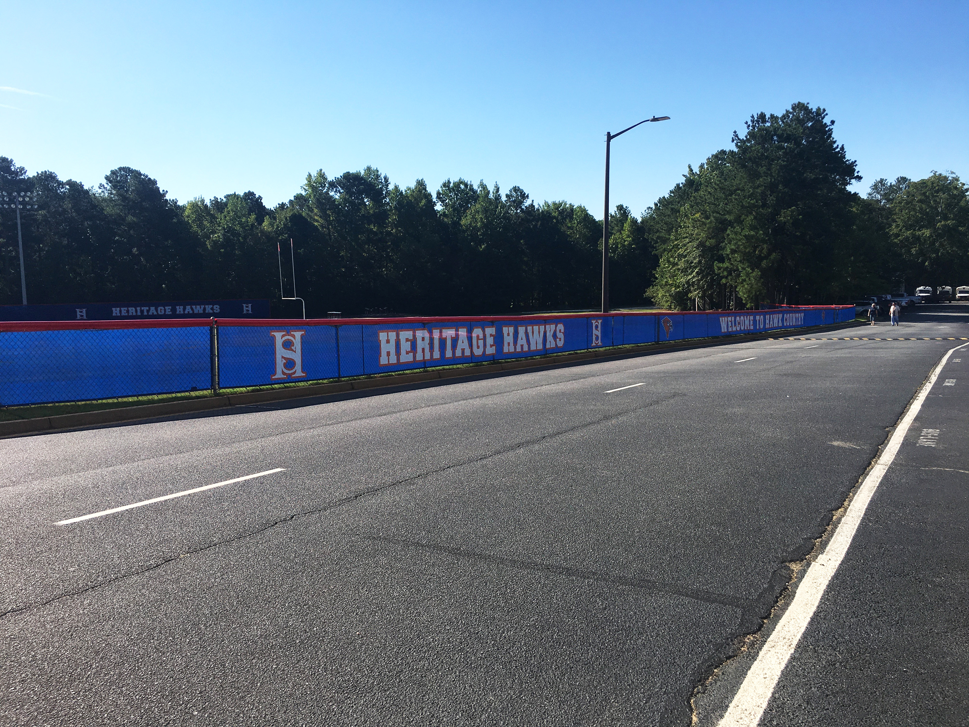 View of fencing with branded windscreen surrounding a football field, separating it from the surrounding area and a nearby road, as seen from the opposite side of the road. The fence is topped with a red fence cap, and the blue windscreen is printed with various logos and text from left to right: an 'HS' monogram logo where the two letters are combined, the words 'HERITAGE HAWKS,' another 'HS' monogram logo, a red cardinal head logo, the phrase 'WELCOME TO HAWK COUNTRY,' and another cardinal head logo. All text and logos are printed in white with red outlines.