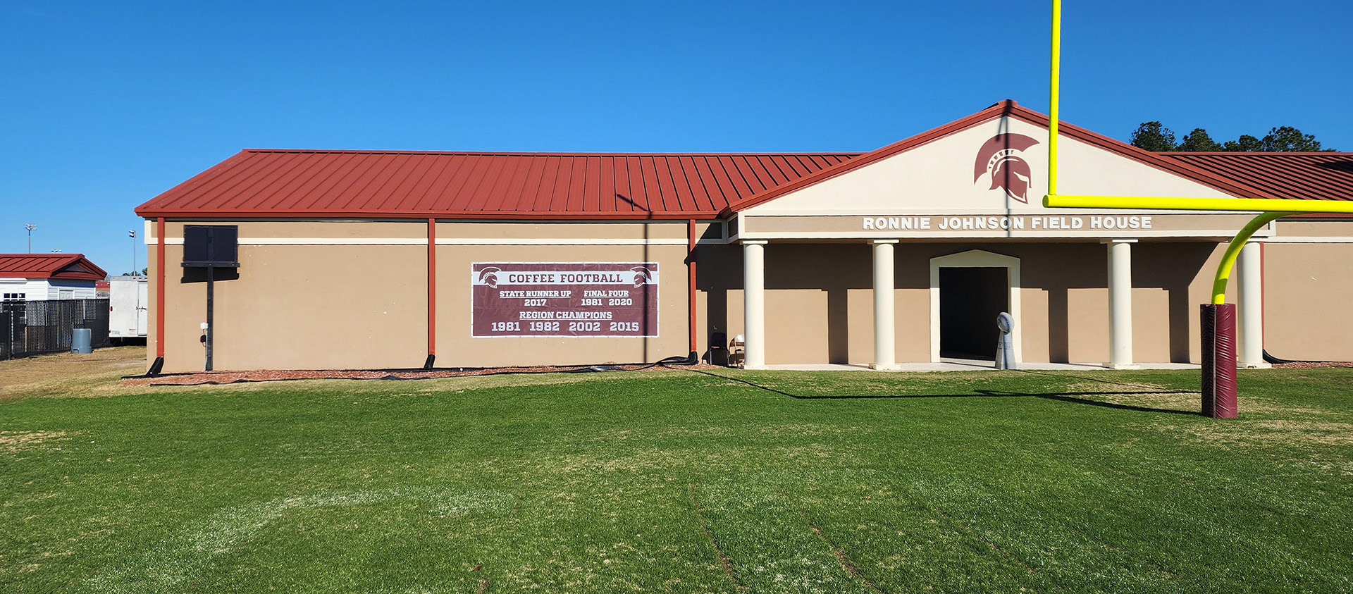 Ronnie Johnson Field House, painted with a Spartan logo above the entrance. On the right side of the building, a windscreen banner displays the years for state runner-up, Final Four, and region champion for 'Coffee Football.' The building is located behind a football goal.