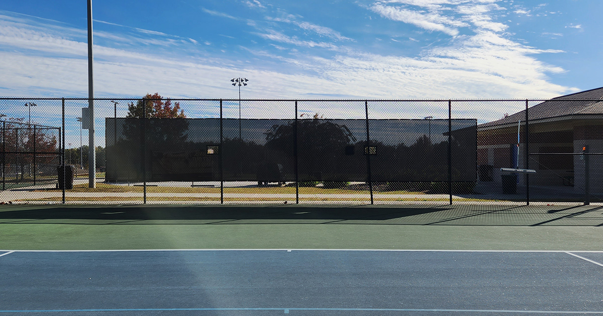 Tennis courts with a fence enclosure, affixed with a black windscreen panel featuring square vents.