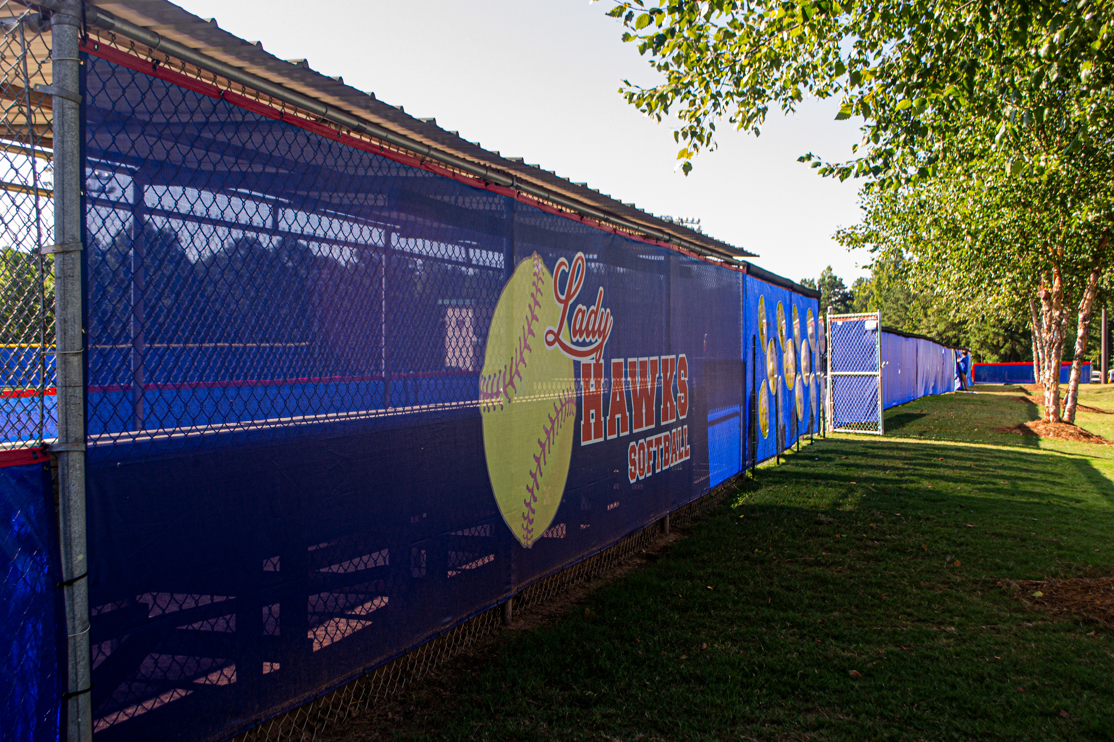 To the left is the back of a dugout and extended fencing with blue windscreen surrounding a baseball field, and to the right is grass and a few trees. The back of the dugout displays red text that reads 'LADY HAWKS SOFTBALL' outlined in white, with a yellow softball with red laces partially in the background. A windscreen panel on the fence to the right of the dugout has several circular images, but they are undecernable due to the angle. The remaining blue windscreen on the fence is unbranded, and all the windscreen has red binding along the top outer edge.