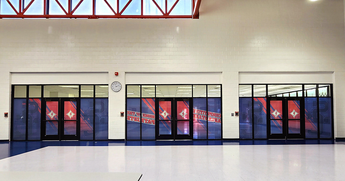 A red, white, and blue window perforation featuring the SC logo, with the text 'Competitive Advantage' and 'Home of the Patriots' displayed on three glass doors inside a school