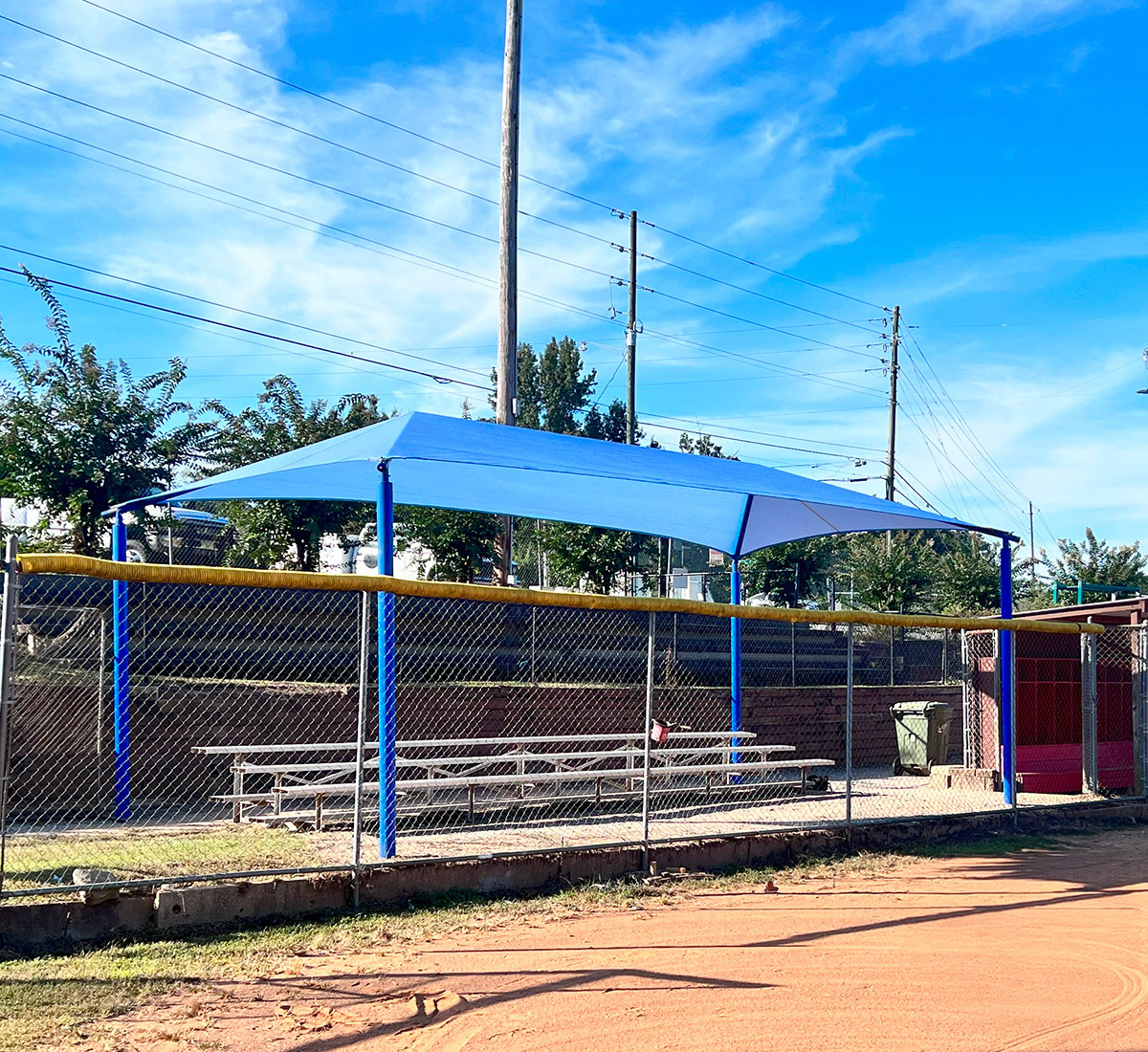 A blue sunshade over a set of bleachers, positioned behind a fence that is located behind a baseball field.