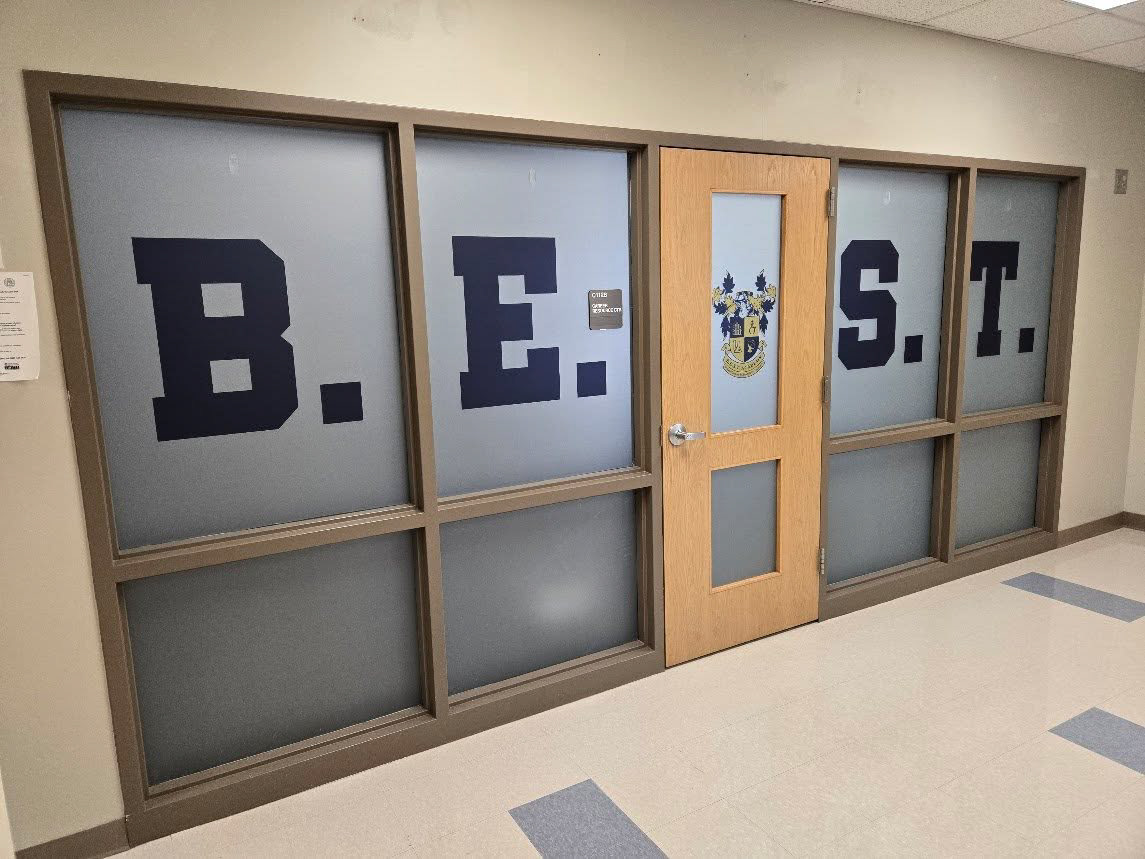 Left-angled view of an interior school doorway showing a windowed door and adjacent windows with custom perforated graphics. Navy blue letters and a yellow and navy blue school crest span the top windows, with frosted window areas below.
