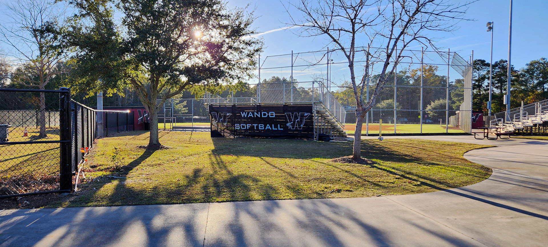 A black windscreen on the backside of metal bleachers near a baseball field. The windscreen features the text 'Wando Baseball' in white, with the 'W' logos on both sides. Each 'W' logo consists of a large black 'W' outlined in burgundy and white, with an arrow weaved through the letter.