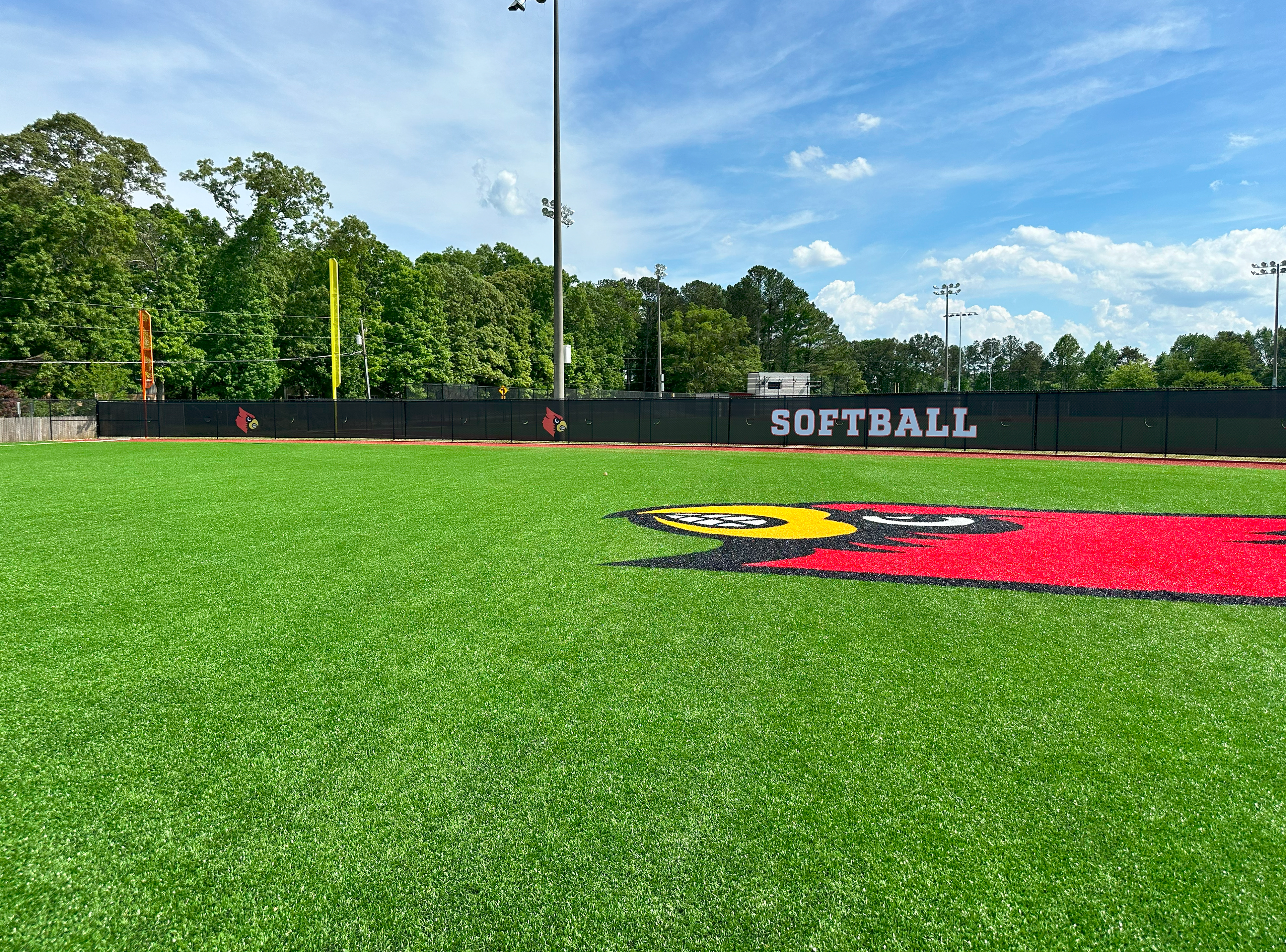 A black windscreen printed with two red cardinal head logos and white text outlined in red that reads 'SOFTBALL,' attached to a fence separating two fields. On the visible field is an image of a red cardinal head logo.