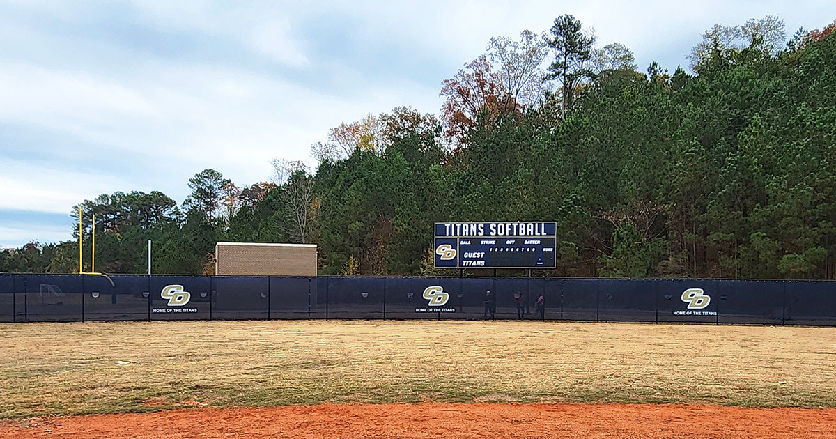 Blue fence windscreen around a baseball field, displaying a repeating 'CD' monogram logo in beige outlined in white and the words 'Home of the Titans' three times. The fence is positioned in front of a scoreboard featuring matching branding.