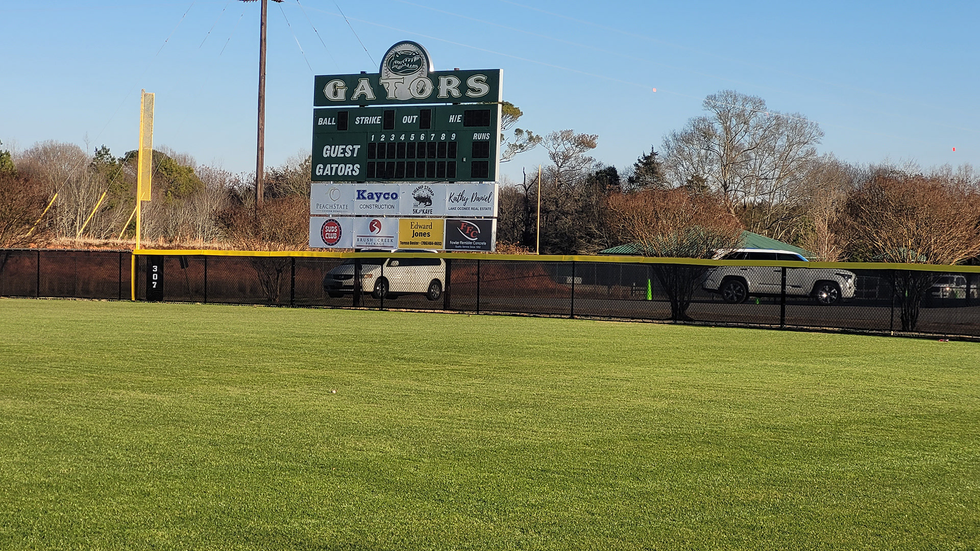 fence surrounding baseball field with black fence windscreen printed with white text reading 'gatewood baseball' and yellow D-style safety top cap, and a black windscreen field maker banner printed with white numbers.