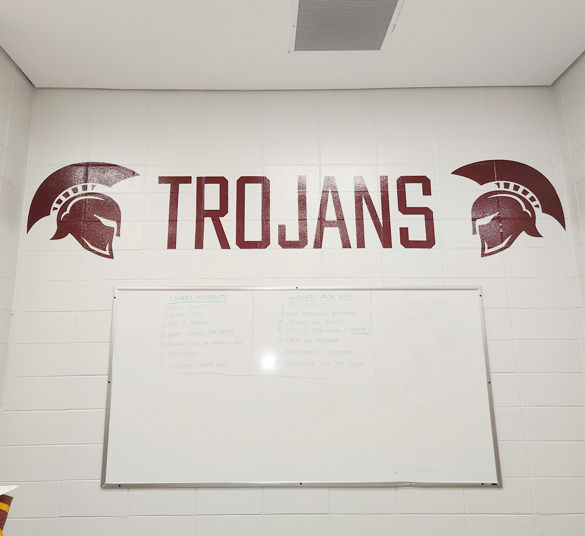 Adhesive vinyl cut burgundy lettering on a painted white brick wall reading "TROJANS" positioned between two images of a Trojan helmets, and above a white board.