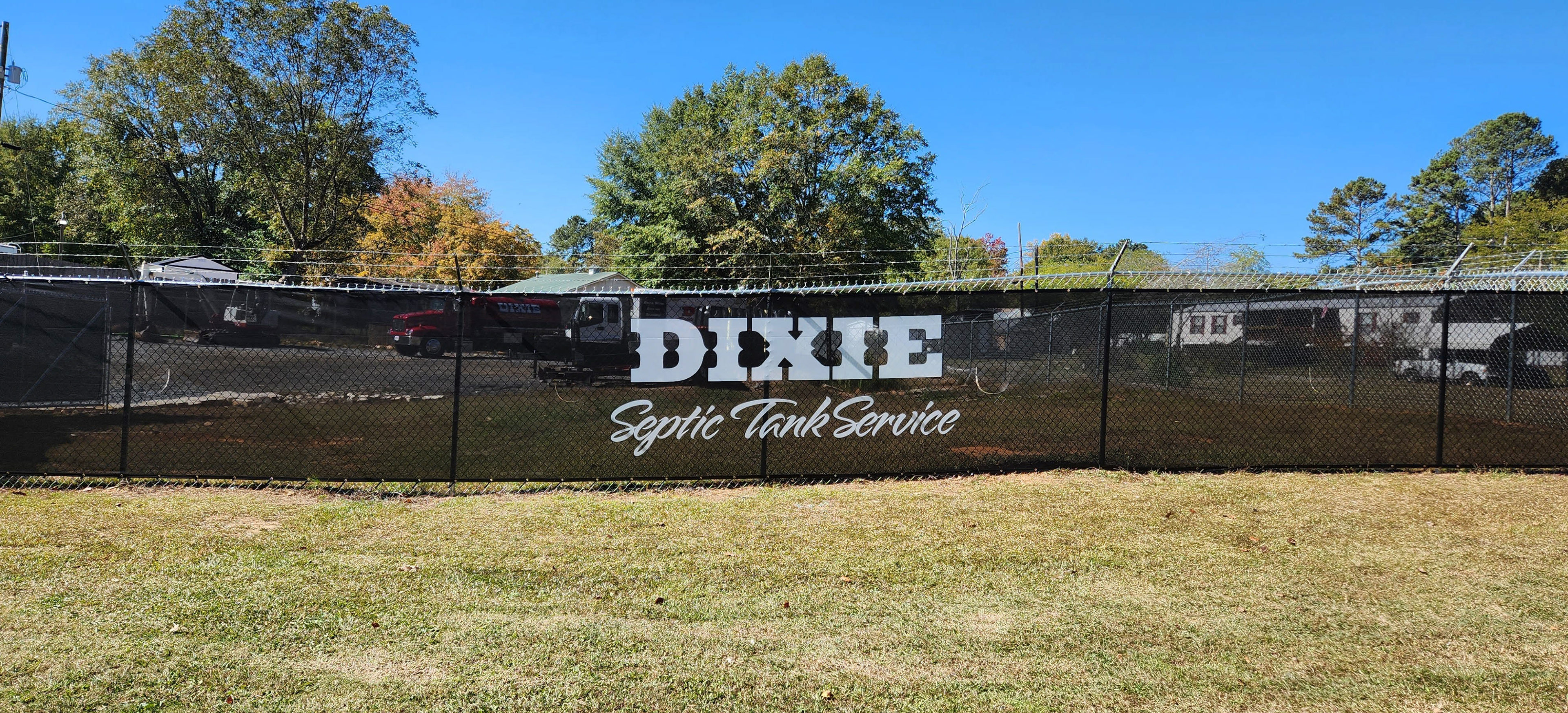 Black windscreen printed with the words 'DIXIE Septic Tank Service' in white, affixed to a fence around a business lot.