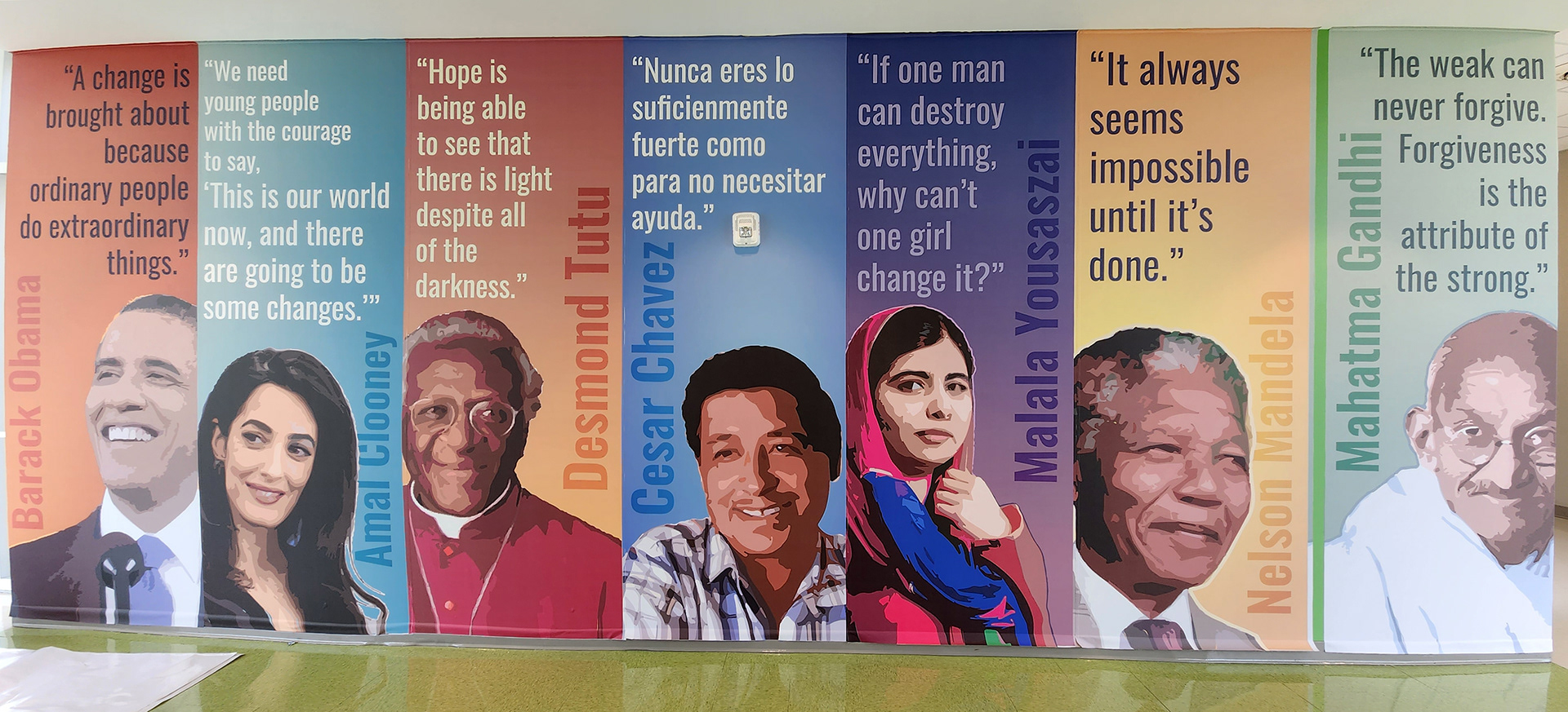 A floor-to-ceiling adhesive vinyl wall mural in a hallway featuring quotes and images of various individuals, each section has a different colored background for their rectangular space. From left to right: The first section has navy text on an orange background reading 'A change is brought about because ordinary people do extraordinary things' above an image of Barack Obama with his name in darker orange. The second section has white text on a light blue background reading 'We need young people with the courage to say, This is our world now, and there are going to be some changes' above an image of Amal Clooney with her name in darker blue. The third section has yellow text on an orange background reading 'Hope is being able to see that there is light despite all of the darkness' above an image of Desmond Tutu with his name in darker orange. The fourth section has white text on a blue background reading 'Nunca eres lo suficientemente fuerte como para no necesitar ayuda' above an image of Cesar Chavez with his name in darker blue. The fifth section has light purple text on a purple background reading 'If one man can destroy everything, why can't one girl change it?' above an image of Malala Yousafzai with her name in darker purple. The sixth section has black text on a yellow background reading 'It always seems impossible until it's done' above an image of Nelson Mandela with his name in darker yellow-orange. The final section has dark green text on a light green background reading 'The weak can never forgive. Forgiveness is the attribute of the strong' above an image of Mahatma Gandhi with his name in green.