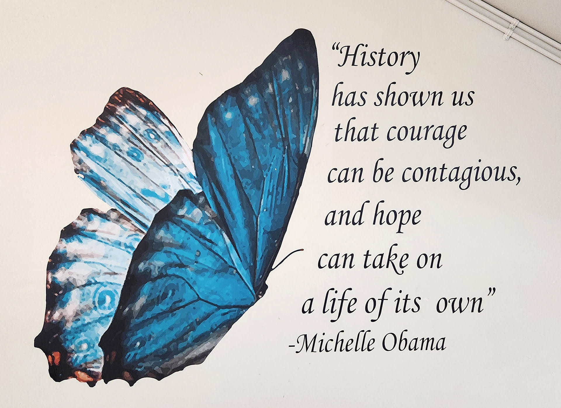 On a wall, a blue butterfly adhesive vinyl decal is positioned to the left of cut adhesive vinyl black lettering that reads 'History has shown us that courage can be contagious, and hope can take on a life of its own' – Michelle Obama. 