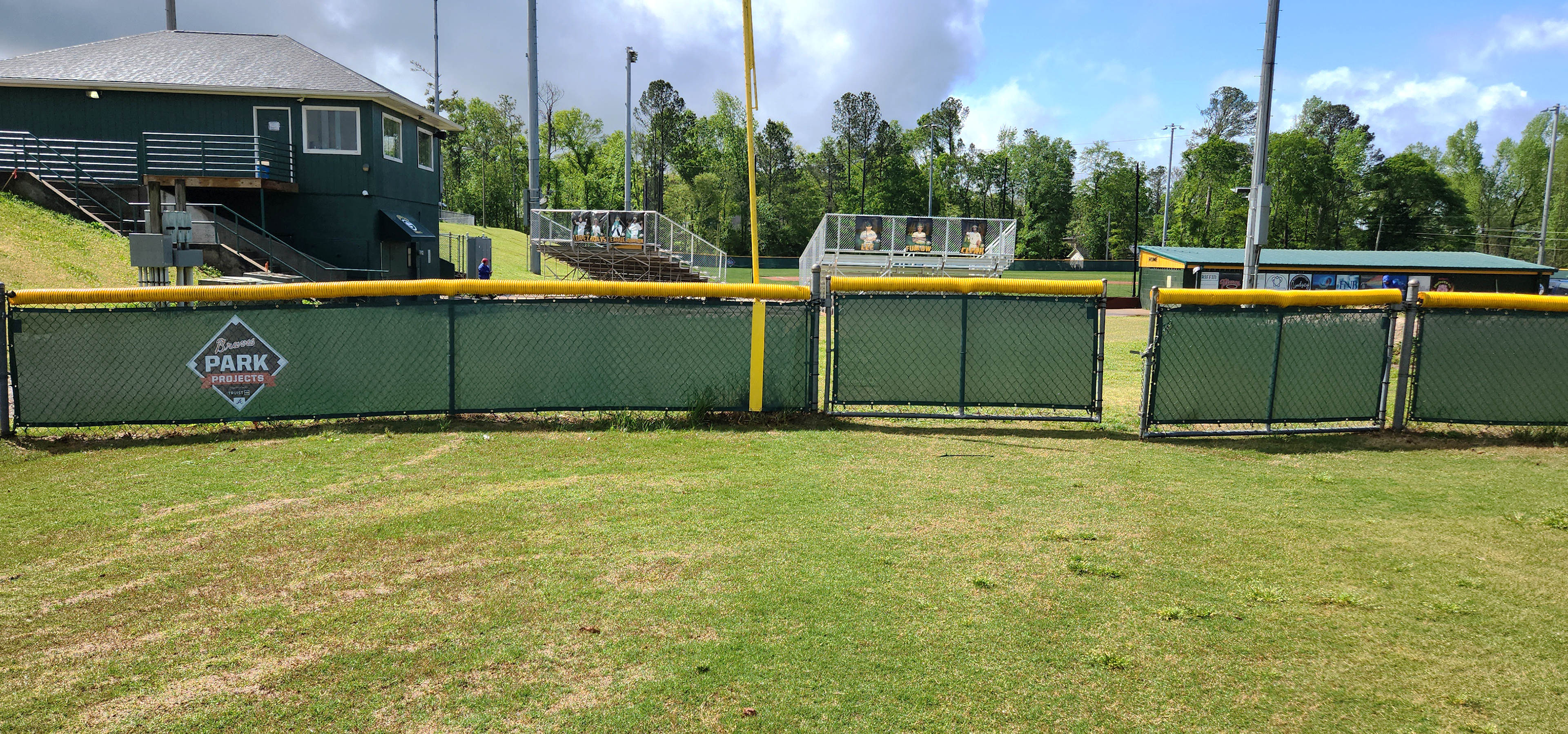 Near shot of dark forest green windscreens that are branded with a navy, red, and white logo reading 'Braves Park Projects' attached to a fence with a yellow corrugated poly cap rail top fence cap.