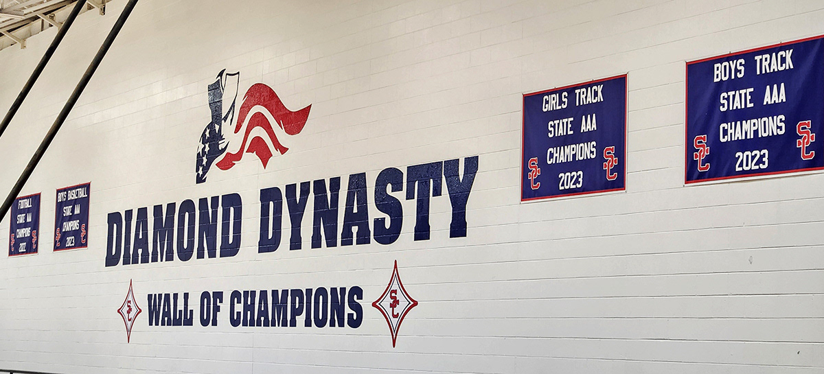 A full-print custom-cut adhesive vinyl decal in navy blue and red featuring an image of a patriot and a flag, displayed on a brick wall inside a school gymnasium. The decal is positioned above the words 'Diamond Dynasty' and 'Wall of Champions,' both in navy blue adhesive vinyl, with two red, four-point SC logo stars on either side.
