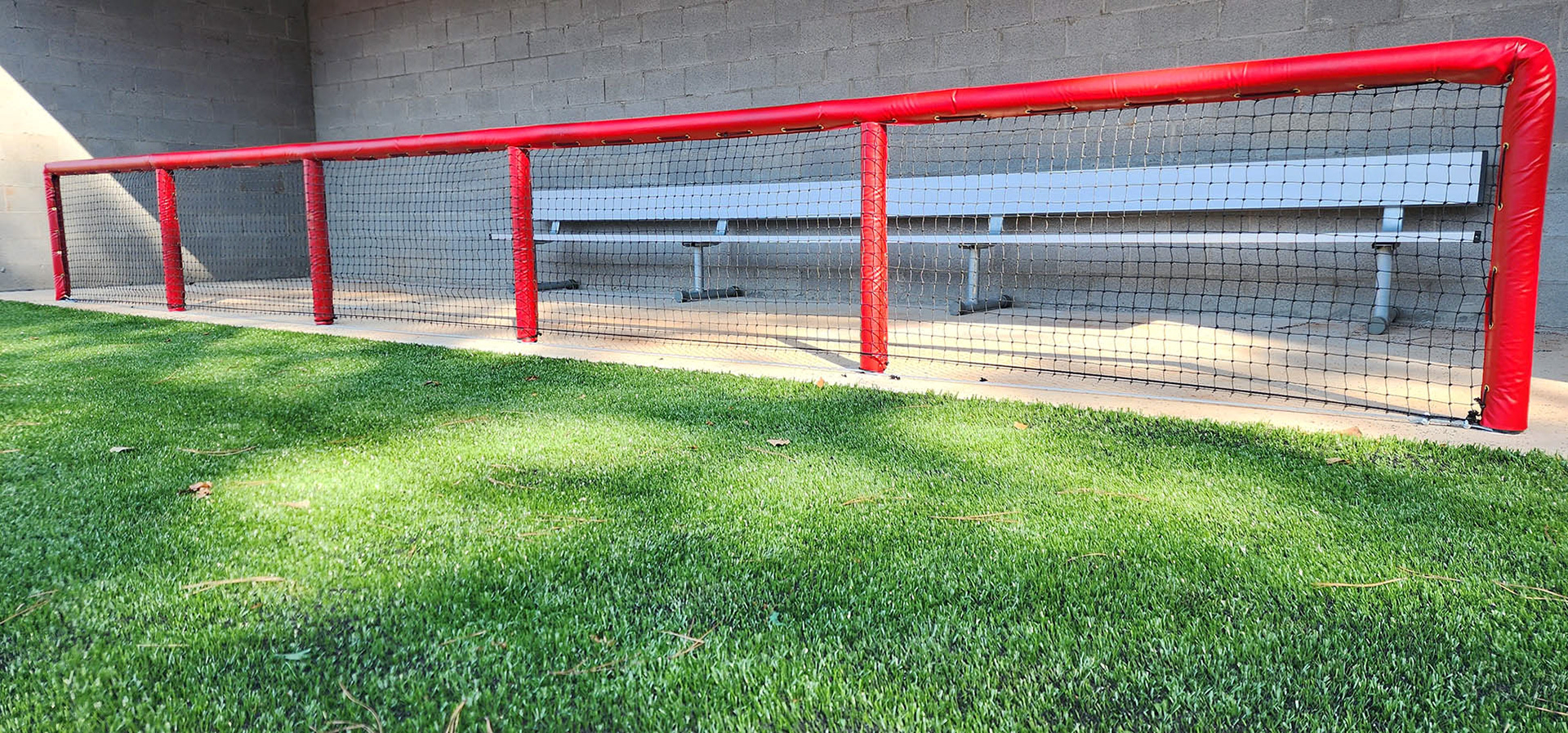 Red vinyl-covered pole padding over the dugout railing, with black sports netting covering the front of the railing to protect players in the dugout. The view is from outside the dugout, with turf in front of the railing and a players' bench behind it.